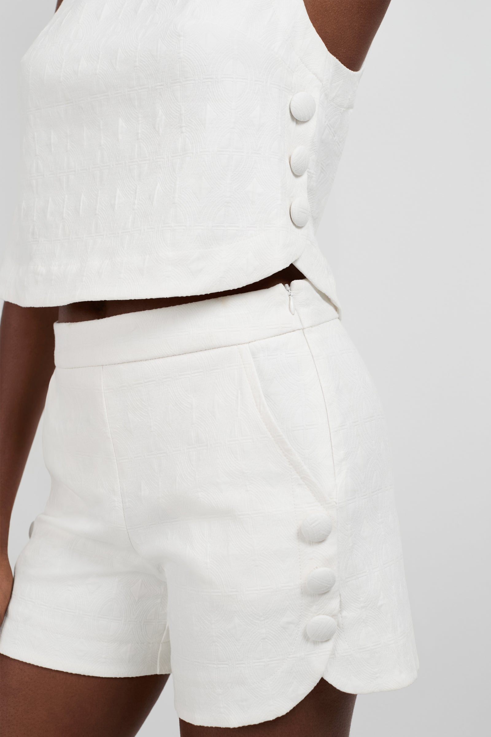 A person wearing the Siara Button Side Top, a white textured sleeveless top with large fabric buttons on the side, photographed from shoulder to mid-thigh against a plain background.