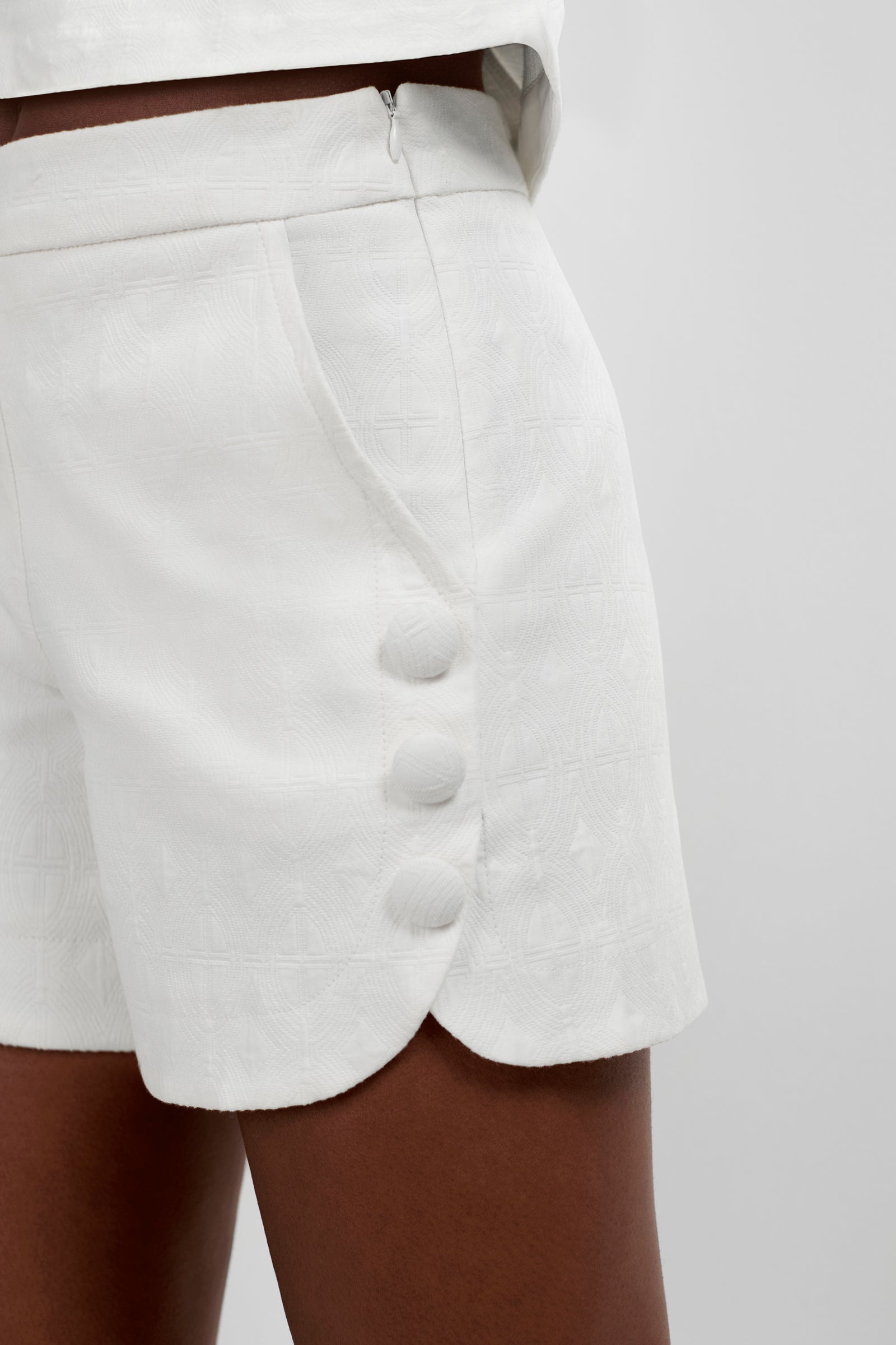 A close-up of a person wearing Duomo Button Side Shorts—white, high-waisted with textured fabric, scalloped hem, three fabric-covered side buttons, side zipper. Their hand rests near the pocket.
