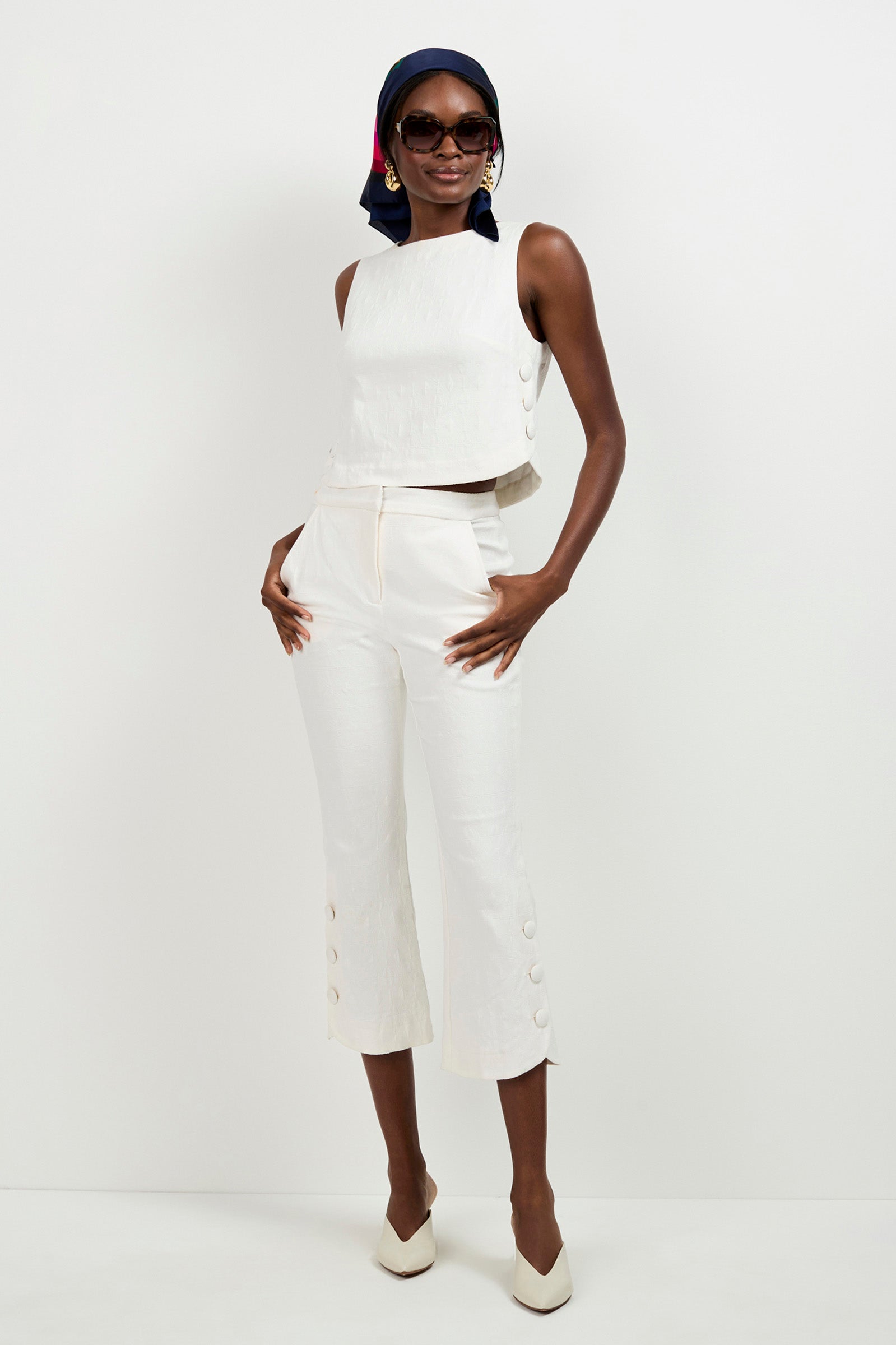A woman poses in white sleeveless crop top and matching Tortona Button Hem Pants, styled with white heels, oversized sunglasses, a navy and red headscarf, and earrings against a plain white background.