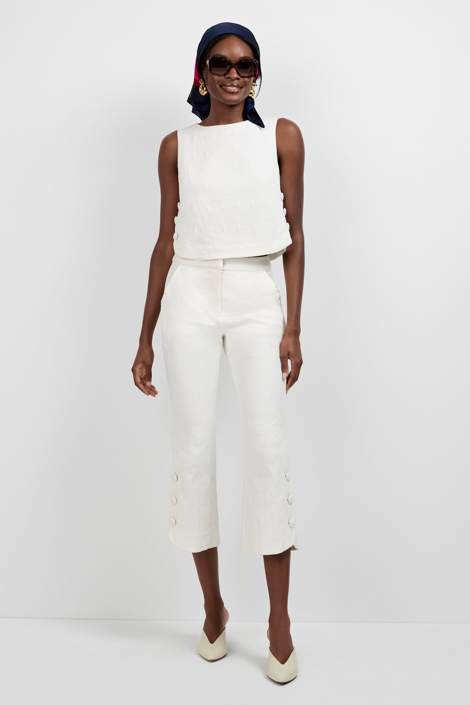 A woman smiles against a plain background wearing Tortona Button Hem Pants, a sleeveless white top, cream heels, sunglasses, gold earrings, and a navy and red headscarf. One hand rests at her side.