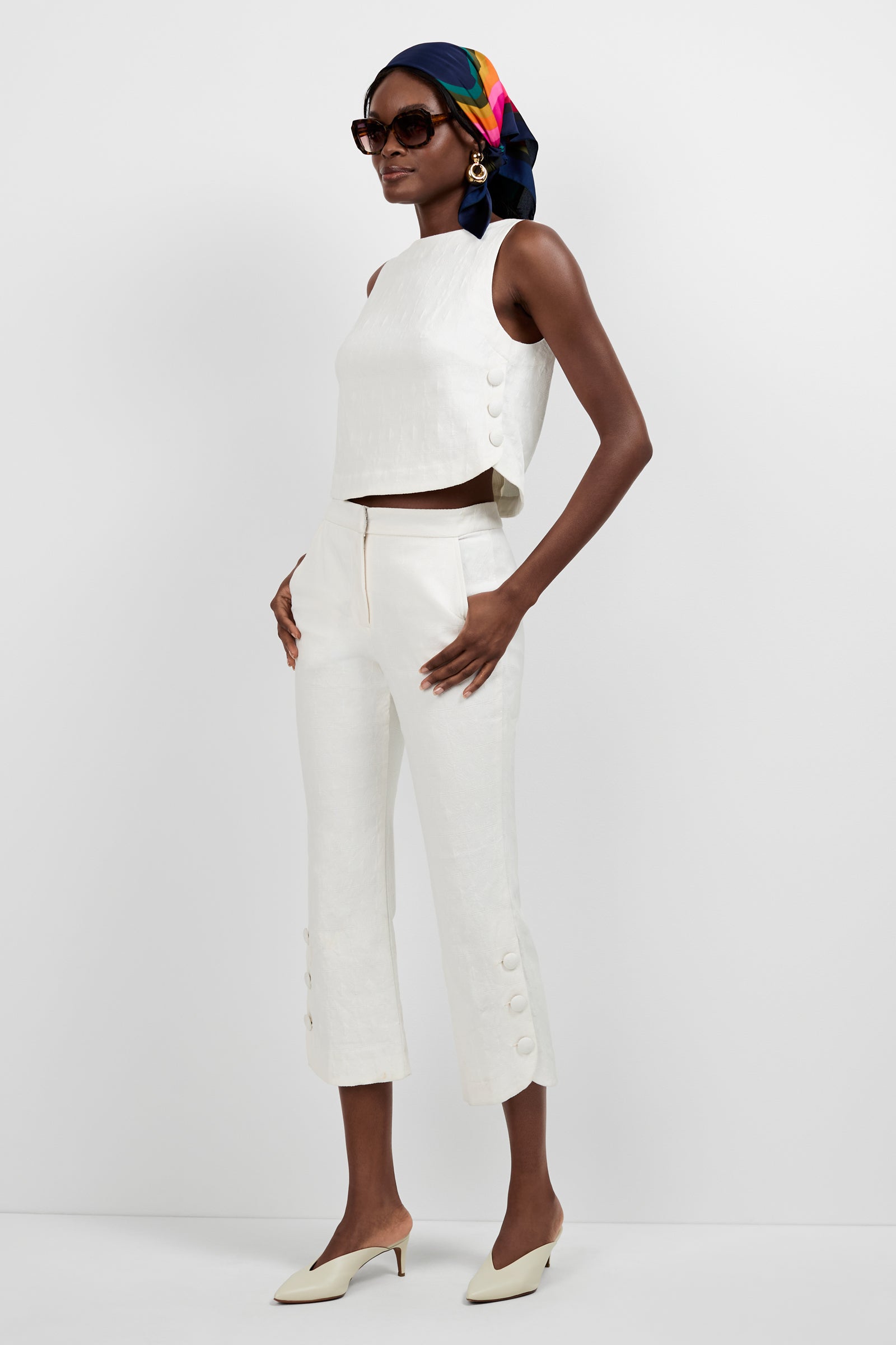 A woman poses in Tortona Button Hem Pants with a matching off-white sleeveless top, cream heels, large sunglasses, statement earrings, and a colorful patterned headscarf.