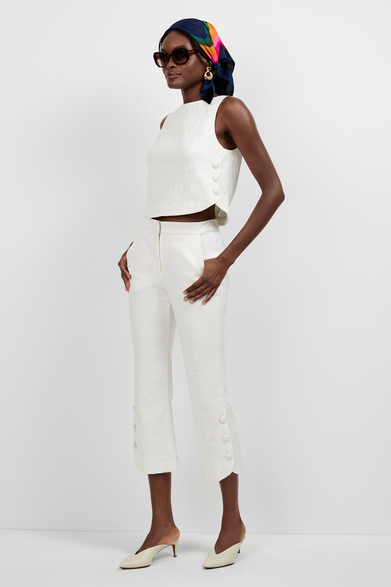 A woman poses in Tortona Button Hem Pants with a matching off-white sleeveless top, cream heels, large sunglasses, statement earrings, and a colorful patterned headscarf.