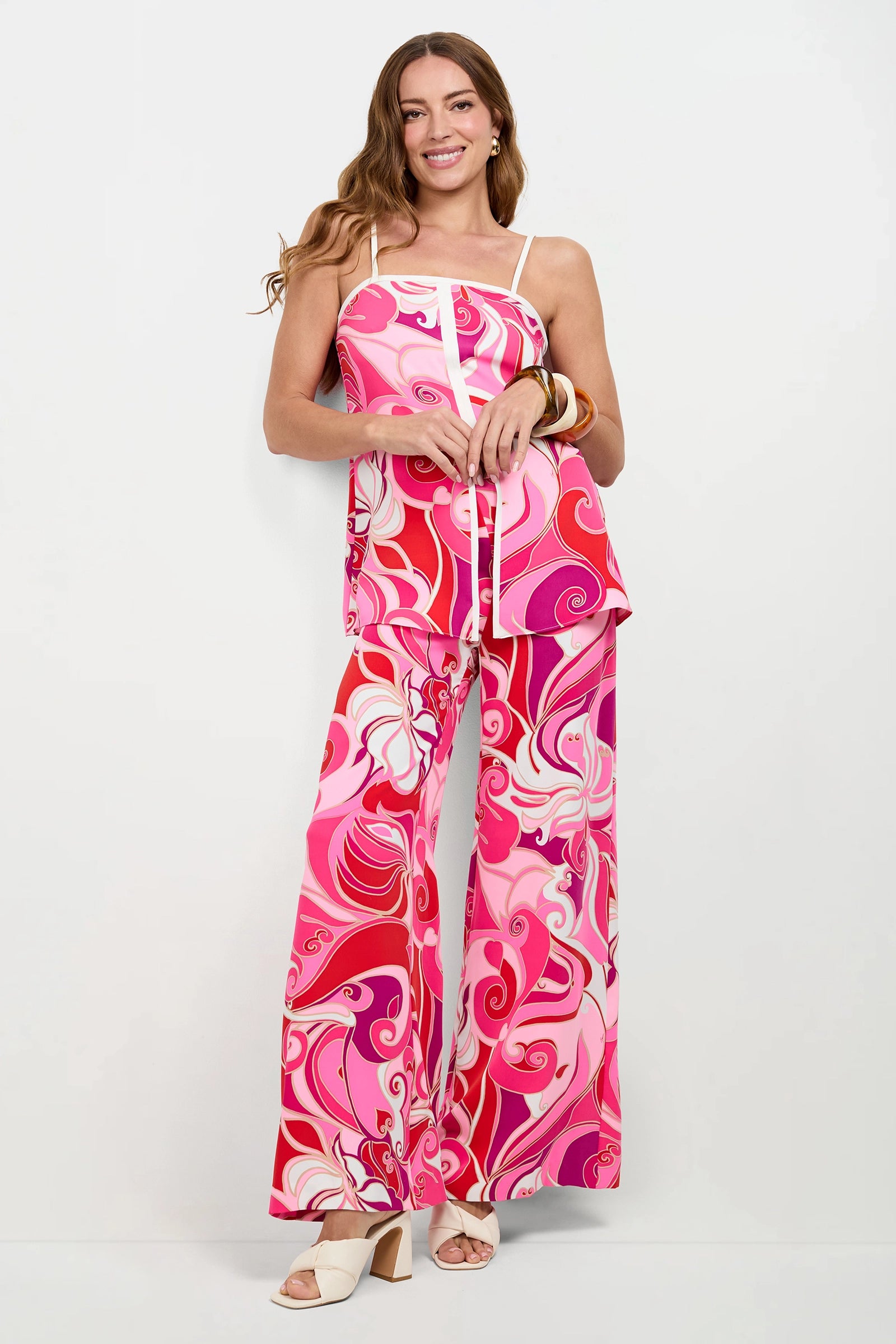 A woman stands smiling in a white room, wearing the Aliza Tunic Top with a pink and red swirl pattern, matching wide-leg pants, white heeled sandals, and large bangles on her wrist.