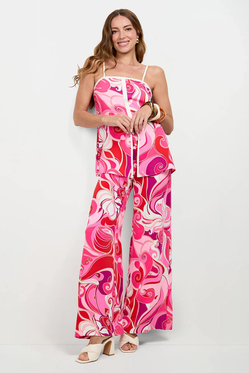 A woman smiles in the Aliza Tunic Top, a colorful pink and red psychedelic print sleeveless piece, paired with matching wide-leg pants. She accessorizes with white heeled sandals and gold bangles against a plain white background.