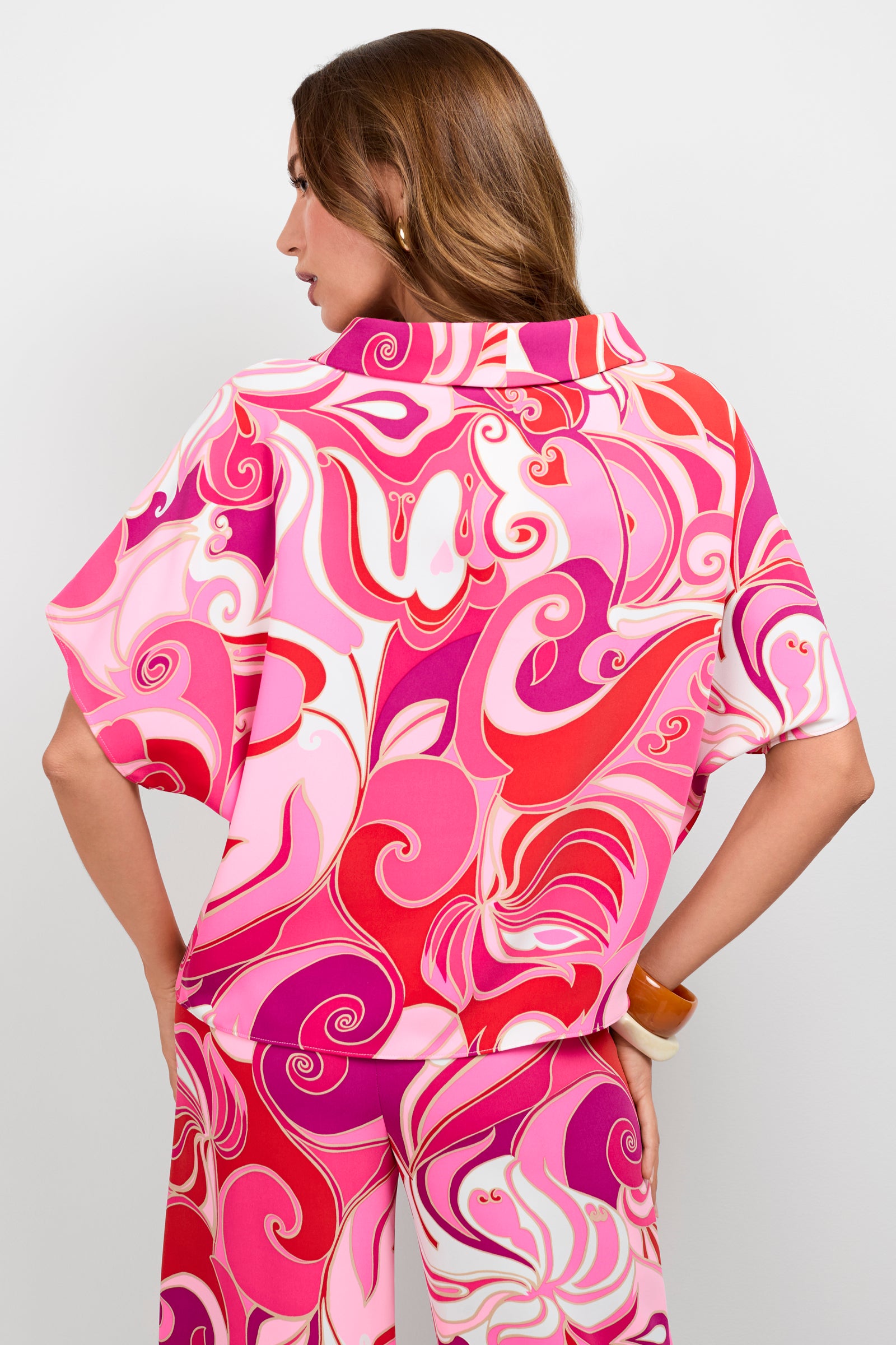 A woman with long brown hair stands facing away, wearing the Honeysuckle Boatneck Top with a bold, swirling pink, red, and white abstract pattern. She's paired it with matching high-waisted pants for a coordinated look.