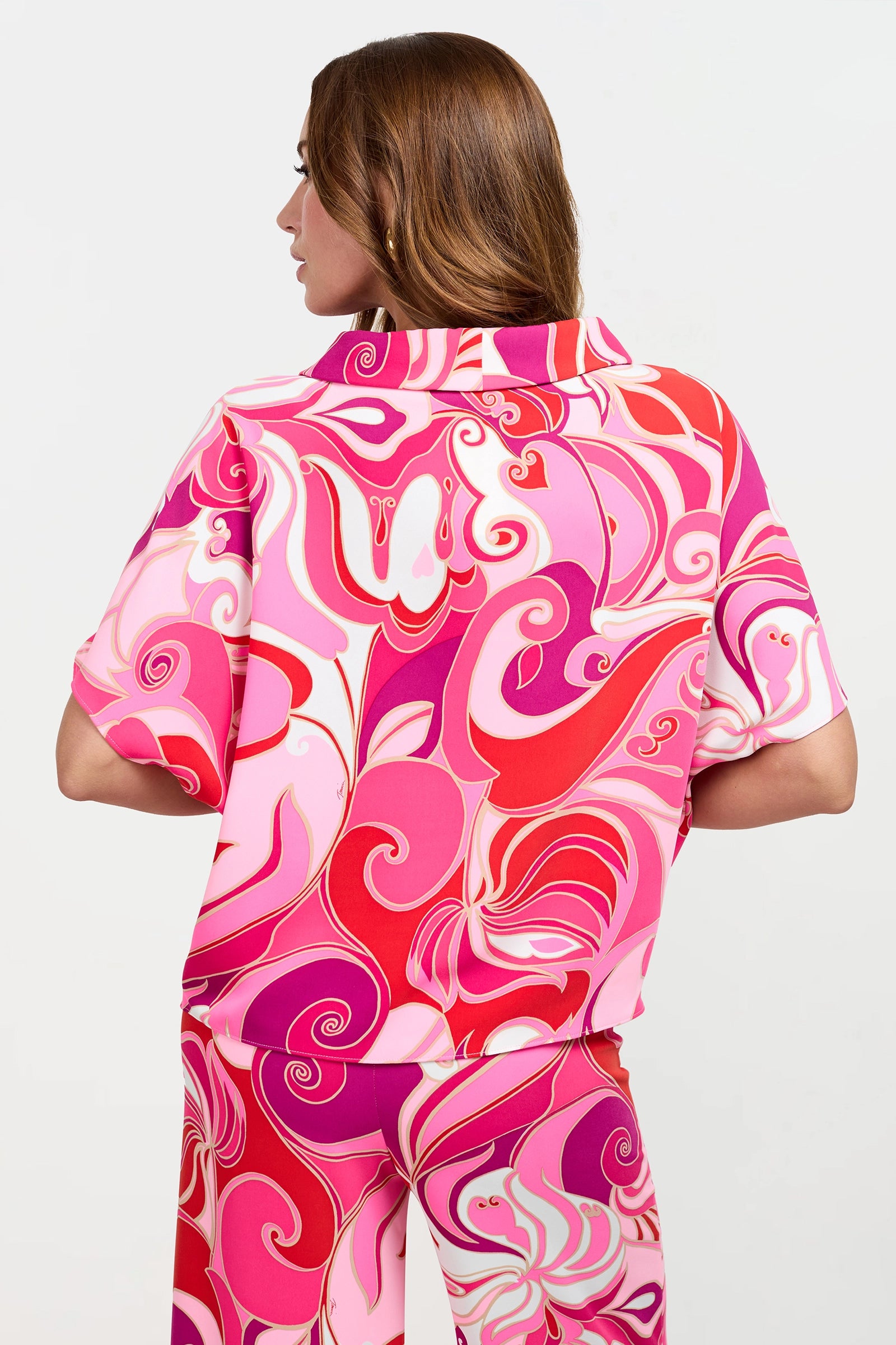A woman with light brown hair is shown from the back, wearing the Honeysuckle Boatneck Top—a colorful, swirling patterned shirt in shades of pink, red, and white—against a plain light background.