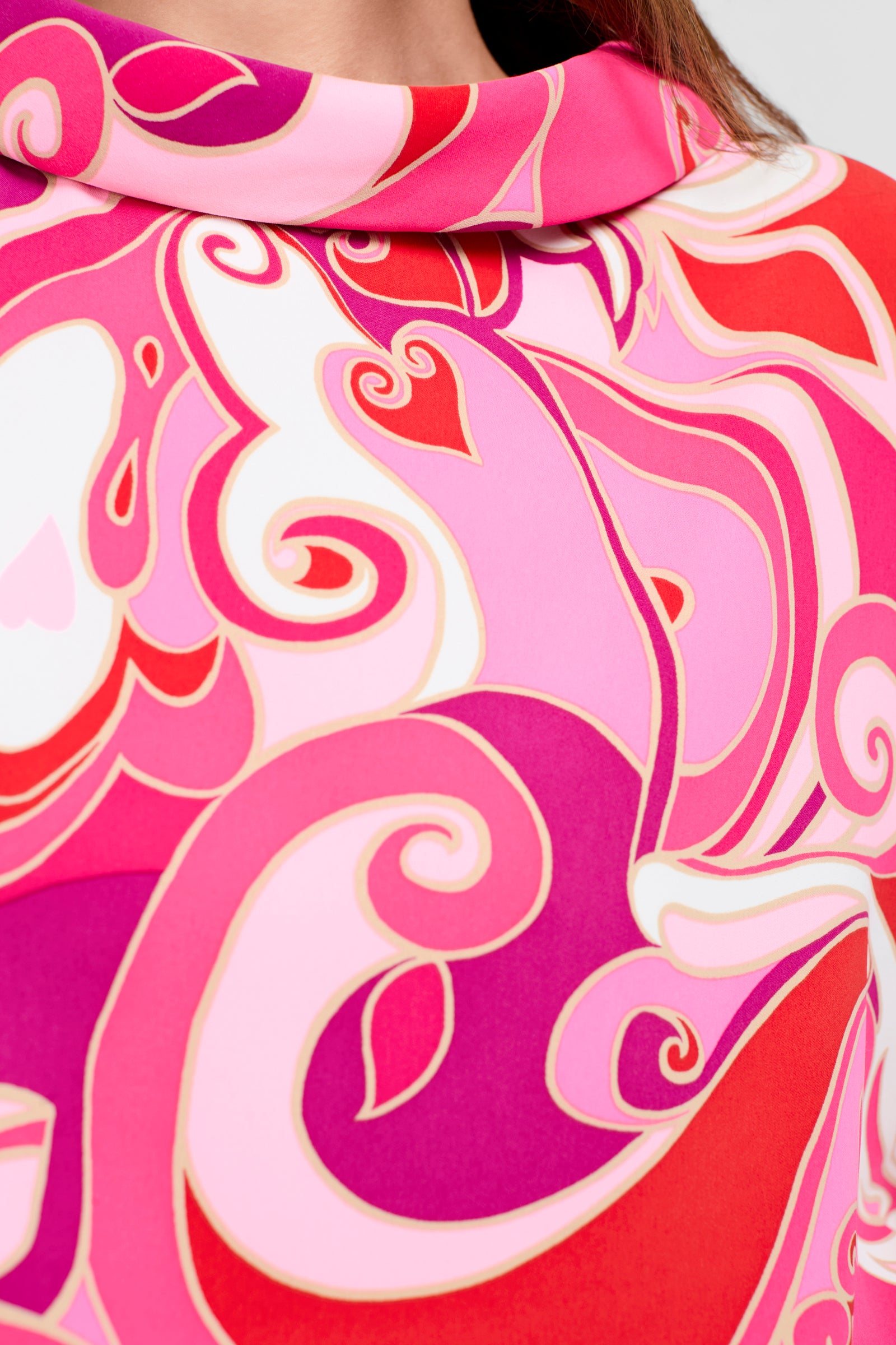 Close-up of the Honeysuckle Boatneck Top, featuring colorful abstract patterns in pink, red, and white with swirling lines. Part of a woman’s neck and brown hair is visible at the top edge.