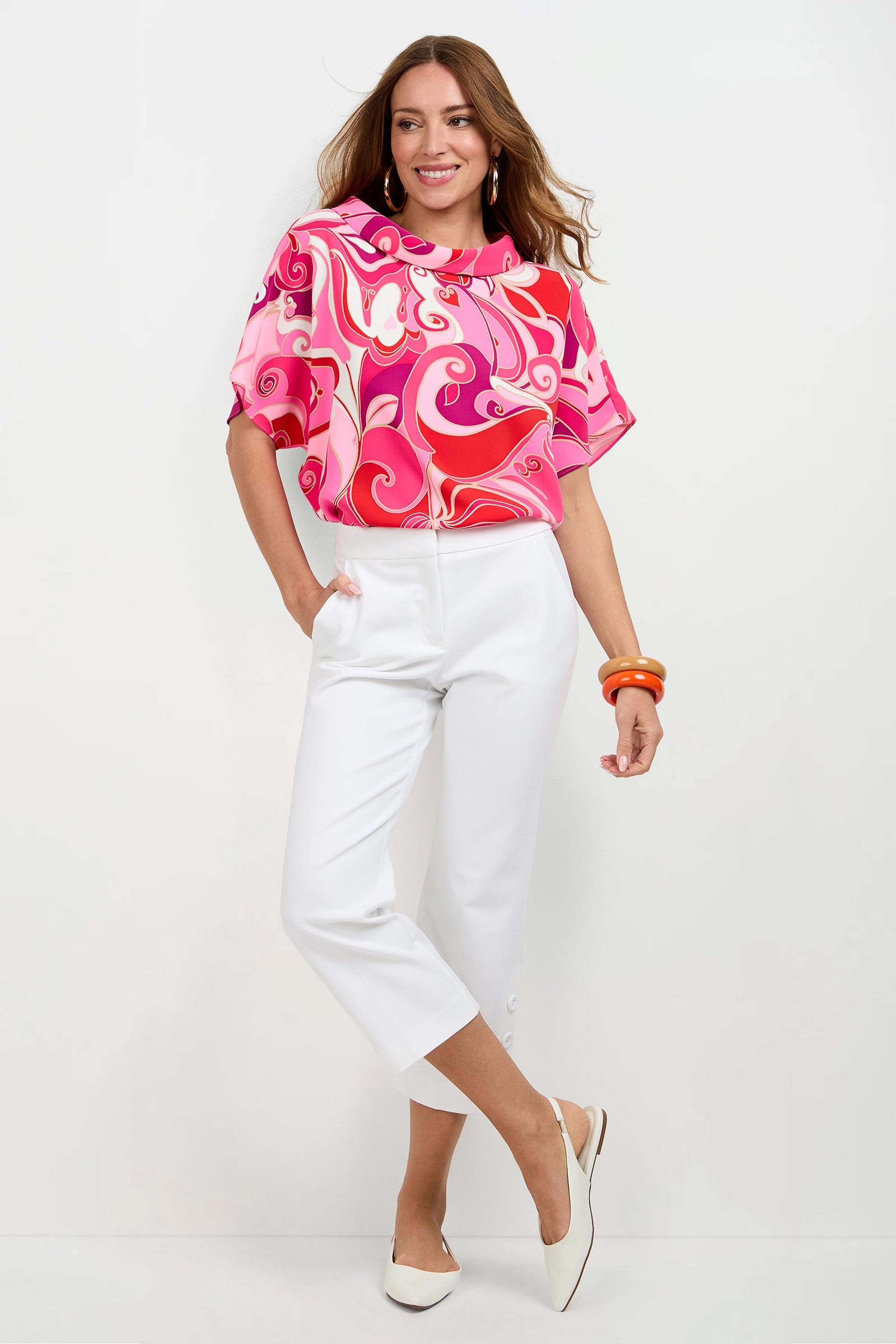 A woman smiles with one hand in her pocket, wearing the Honeysuckle Boatneck Top in vibrant pink and red, white cropped pants, white flats, and orange bangles against a plain white background.