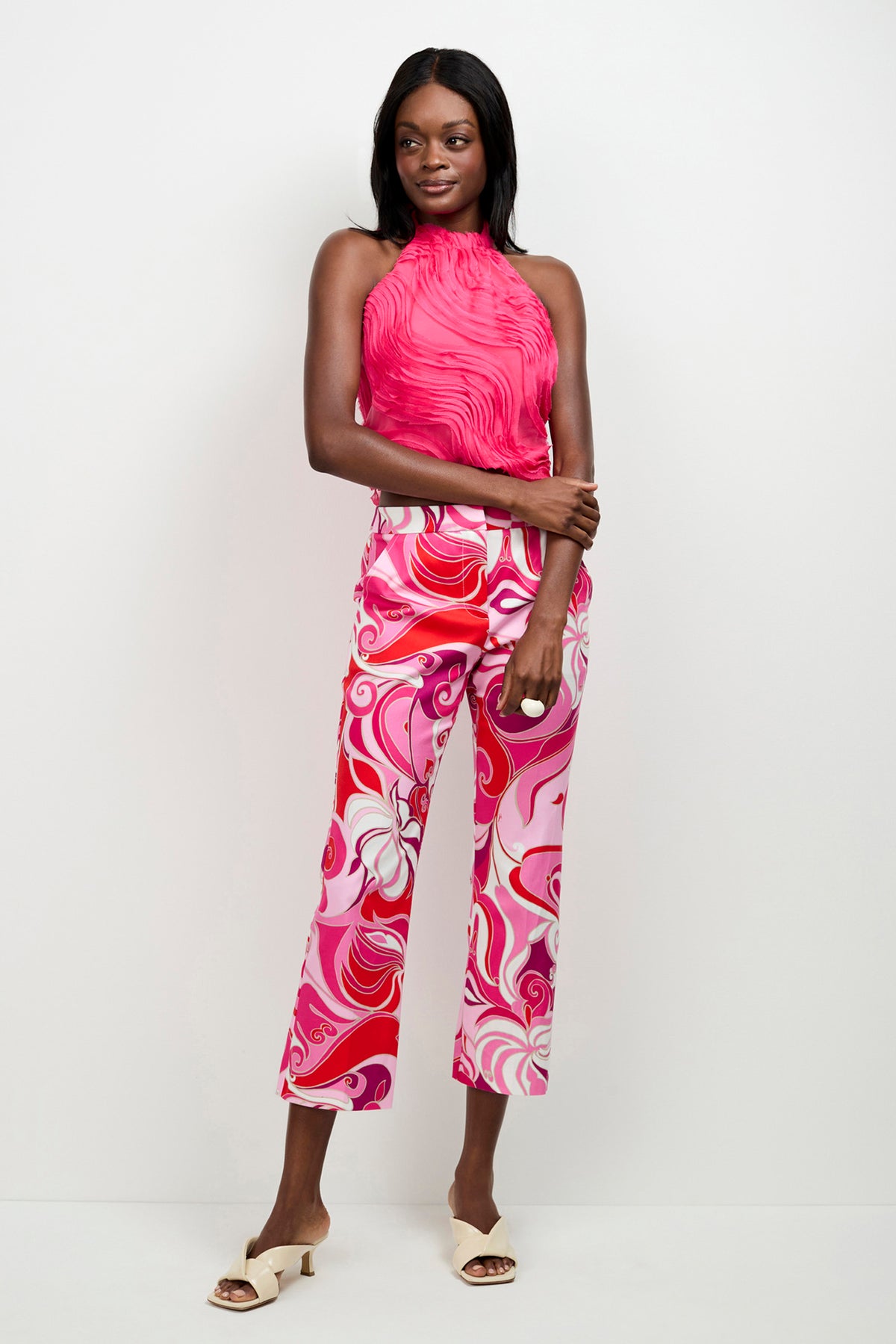 A woman poses against a white wall wearing a sleeveless, bright pink halter top and the Lulu Cropped Pants - Darsana, which feature high-waisted, wide-leg styling with a bold pink, white, and red swirl pattern. She completes the look with white open-toe sandals.