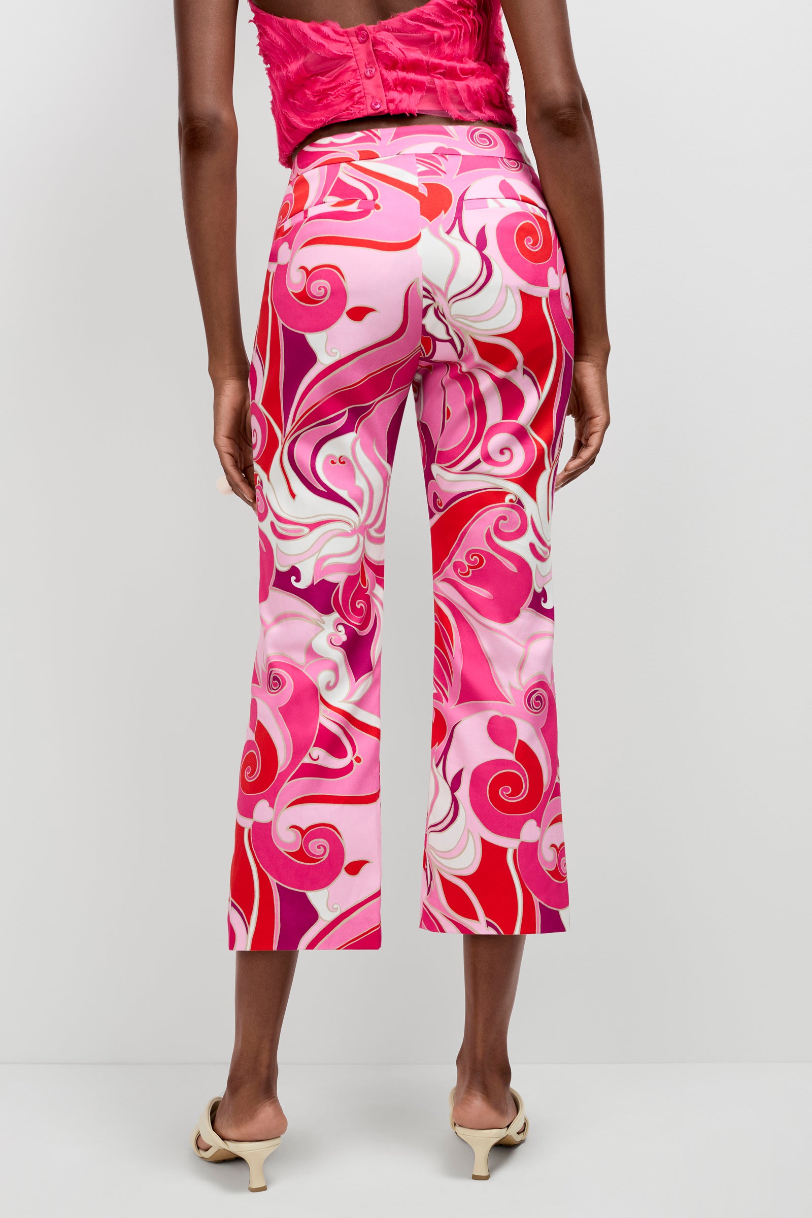 A person wearing the Lulu Cropped Pants - Darsana, featuring a bold swirling pink, red, and white pattern, paired with a pink sleeveless top and cream heeled sandals, shown from the back.