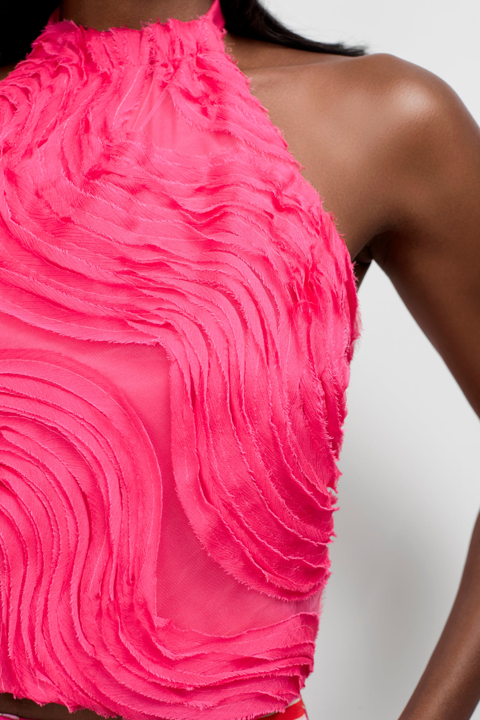 Close-up of a person wearing the Myka Organza Halter Top, a sleeveless, high-neck style made of sheer bright pink fabric with wavy textured patterns and raised detailing. The person's shoulder and part of the chest are visible.