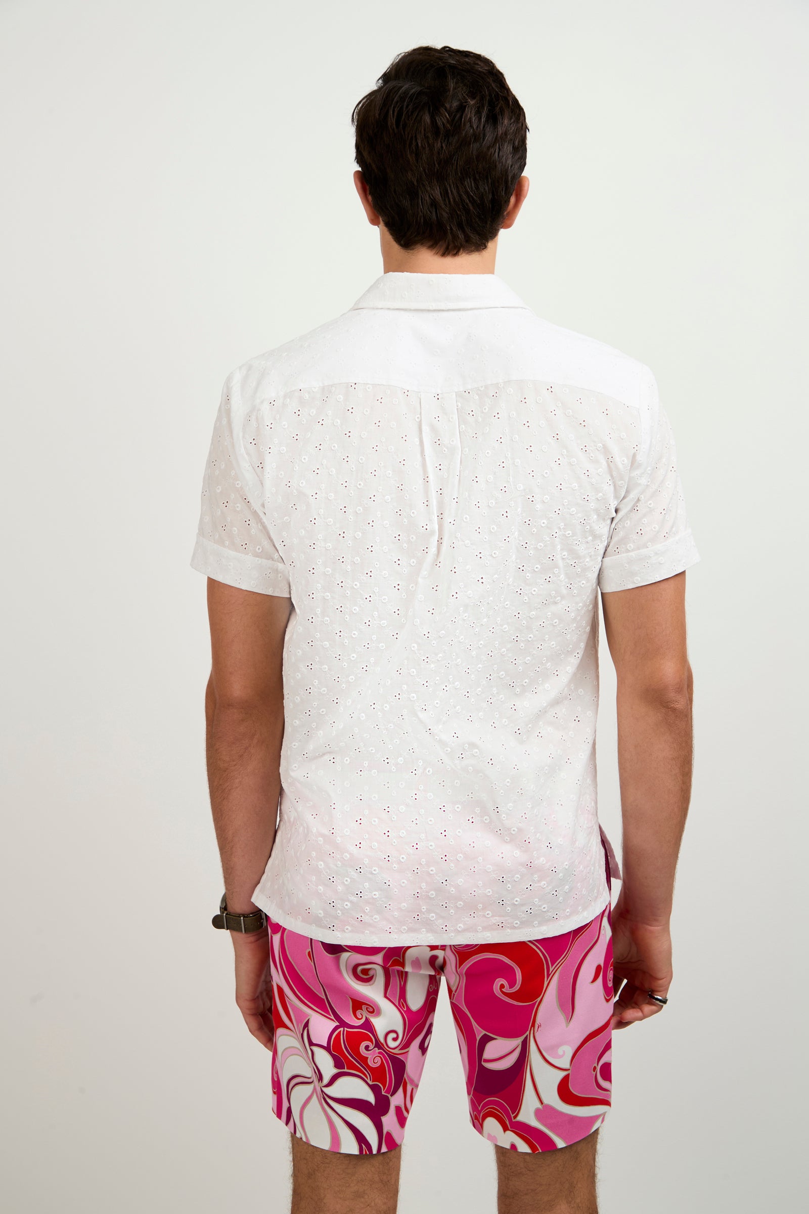 A man faces away from the camera, wearing a white Branson Short-Sleeve Shirt and pink swim trunks with red and white swirling patterns, set against a plain light background.