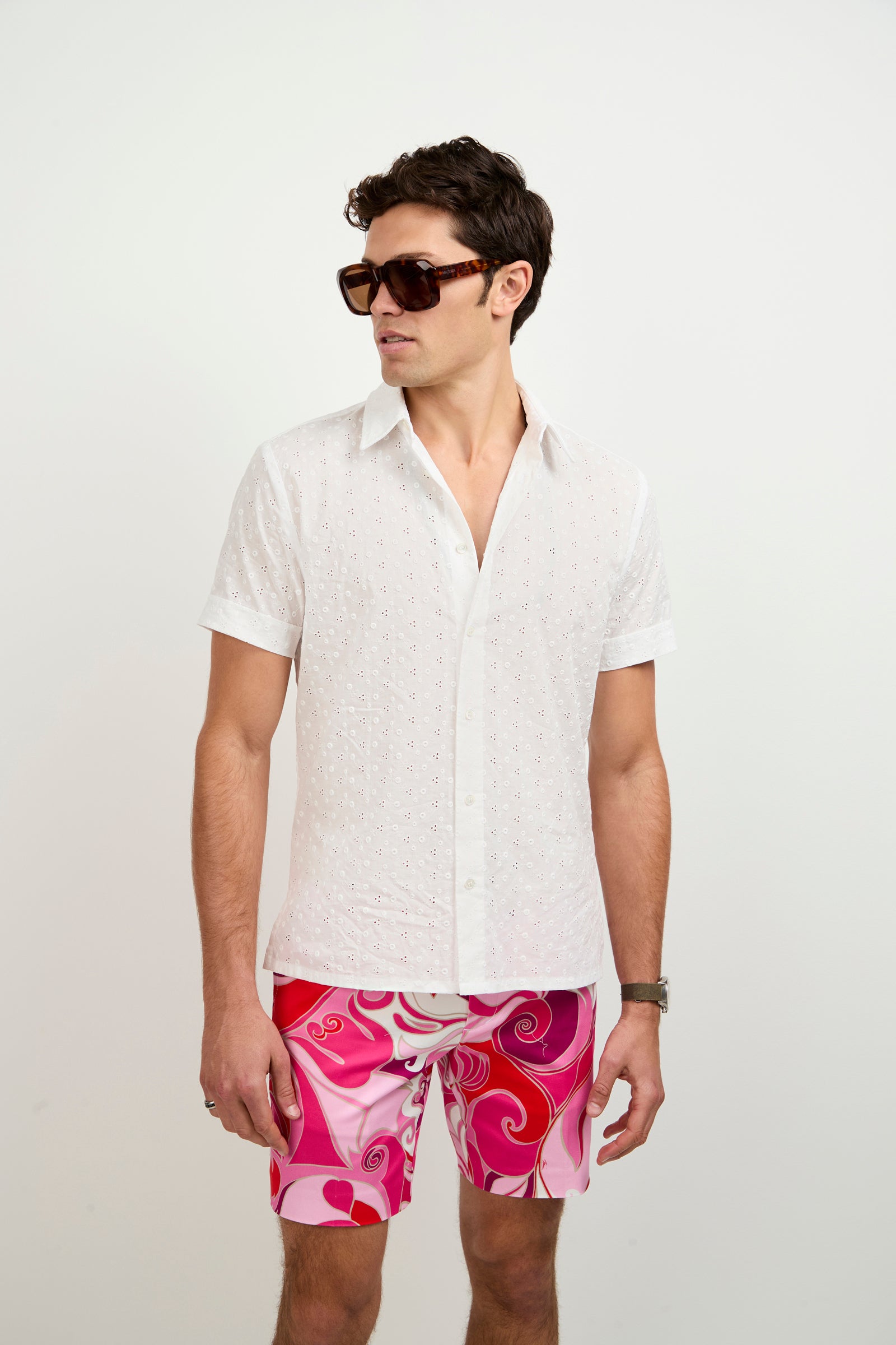 A man in a white Branson Short-Sleeve Shirt, bold pink and red patterned shorts, and large dark sunglasses stands against a plain white background, looking to his left.