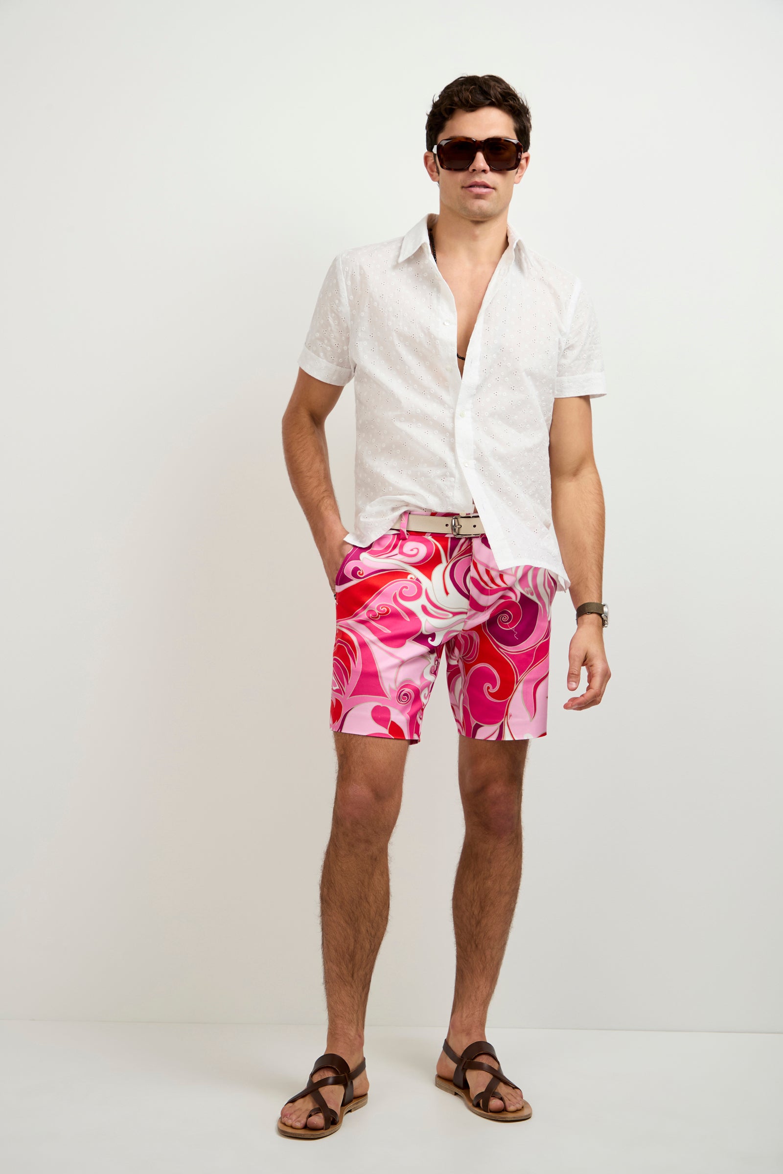 A man in dark sunglasses, the Branson Short-Sleeve Shirt (white), pink and red patterned shorts with a belt, and brown sandals stands against a plain white background.