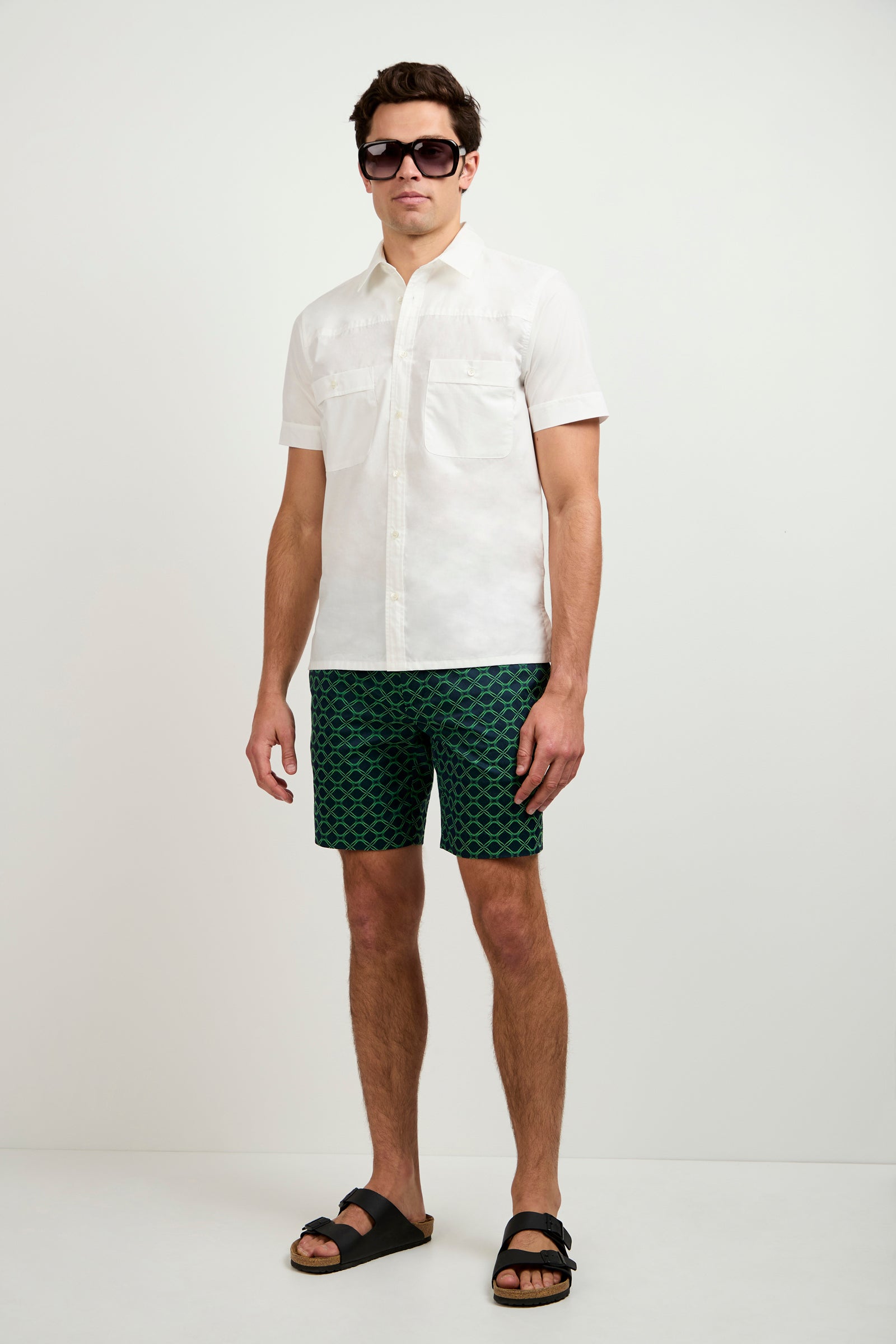 A man in the Will Short-Sleeve Shirt, dark sunglasses, green patterned shorts, and black sandals stands against a plain white background.