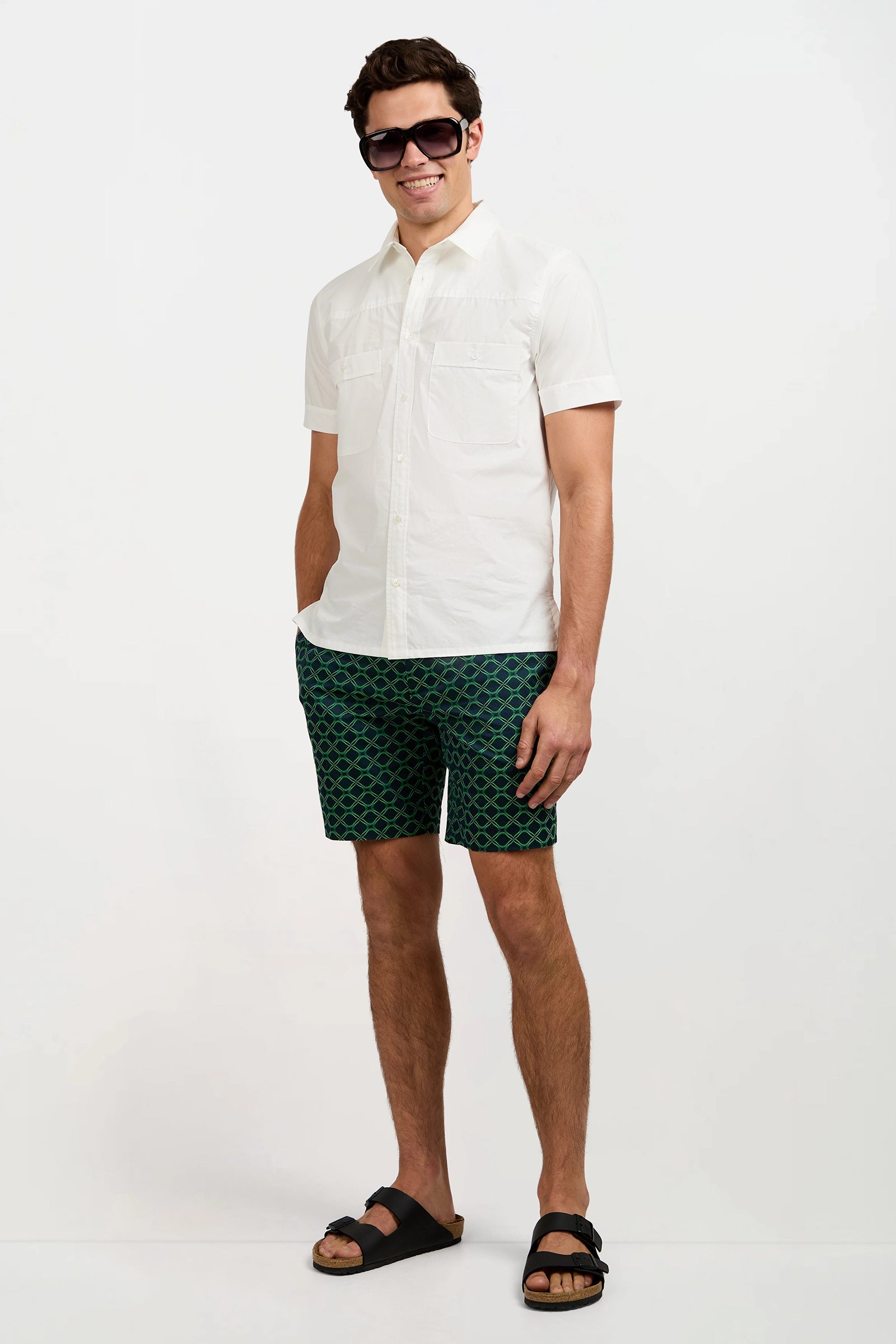A man wearing dark sunglasses, patterned green shorts, black sandals, and the white Will Short-Sleeve Shirt stands smiling with his hands in his pockets against a plain white background.