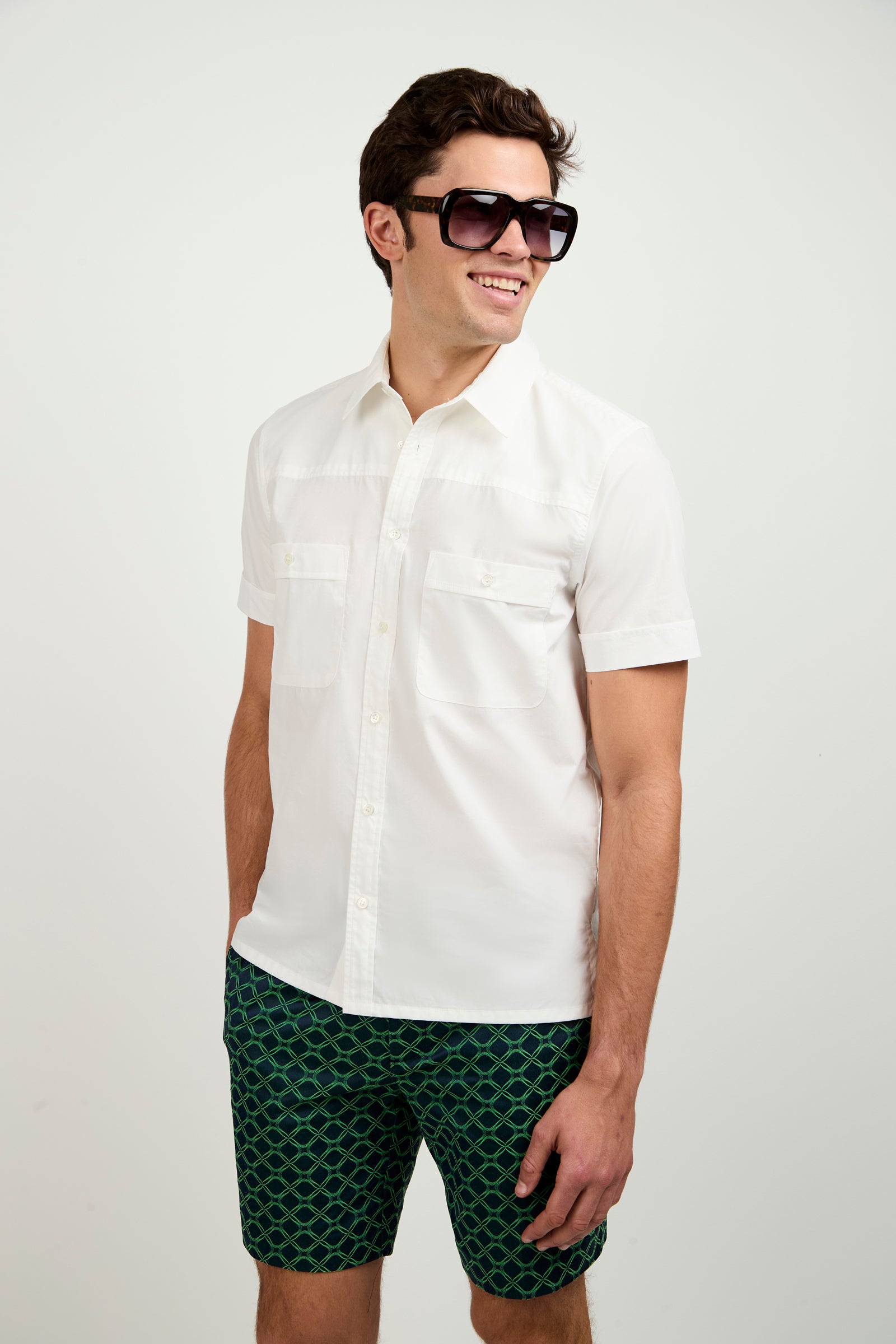 A man wearing the Will Short-Sleeve Shirt, paired with dark sunglasses and green patterned shorts, smiles while looking to the side against a plain white background.