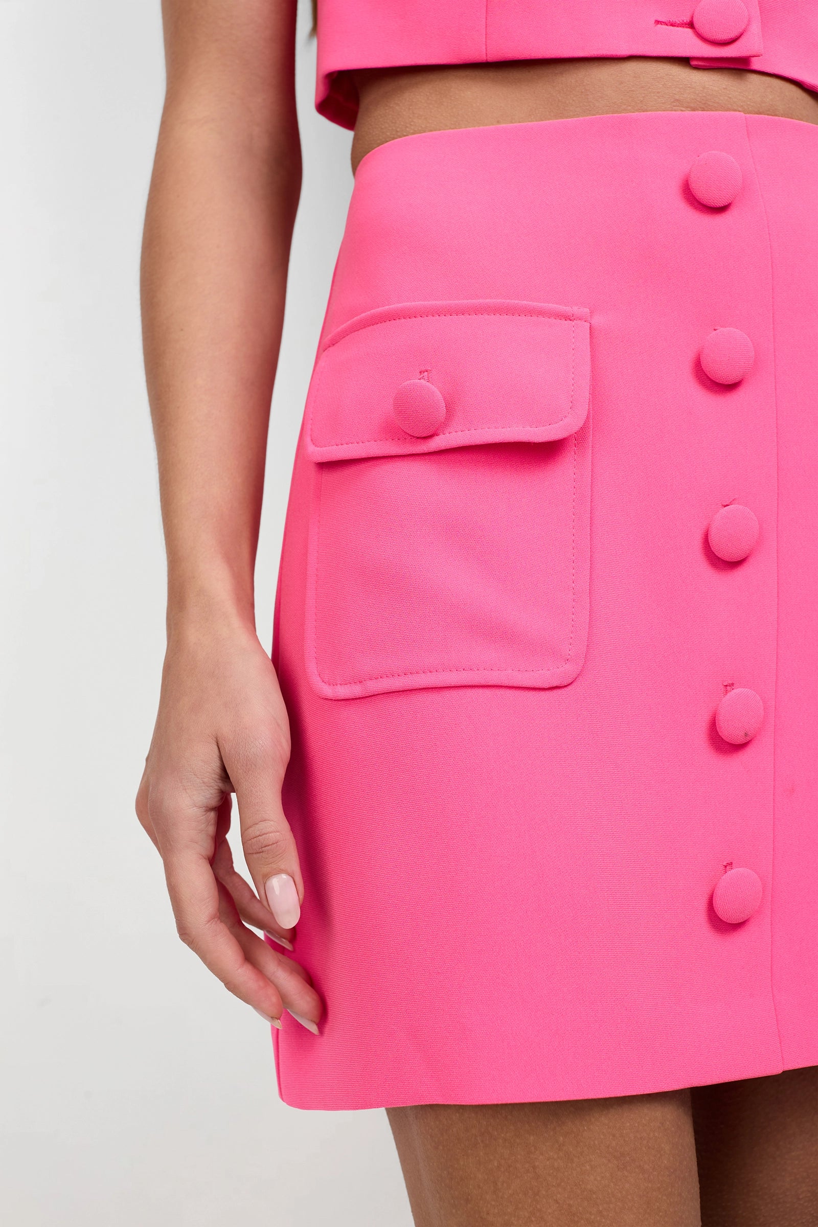 A close-up of a person wearing the Medici Tailored Mini Skirt in bright pink, featuring fabric-covered front buttons and a side flap pocket. The matching pink top is partly visible as their hand rests at their side.