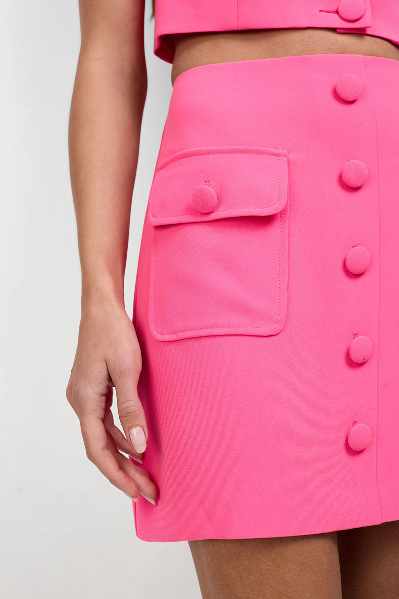 A close-up of a person wearing the Medici Tailored Mini Skirt in bright pink, featuring fabric-covered front buttons and a side flap pocket. The matching pink top is partly visible as their hand rests at their side.