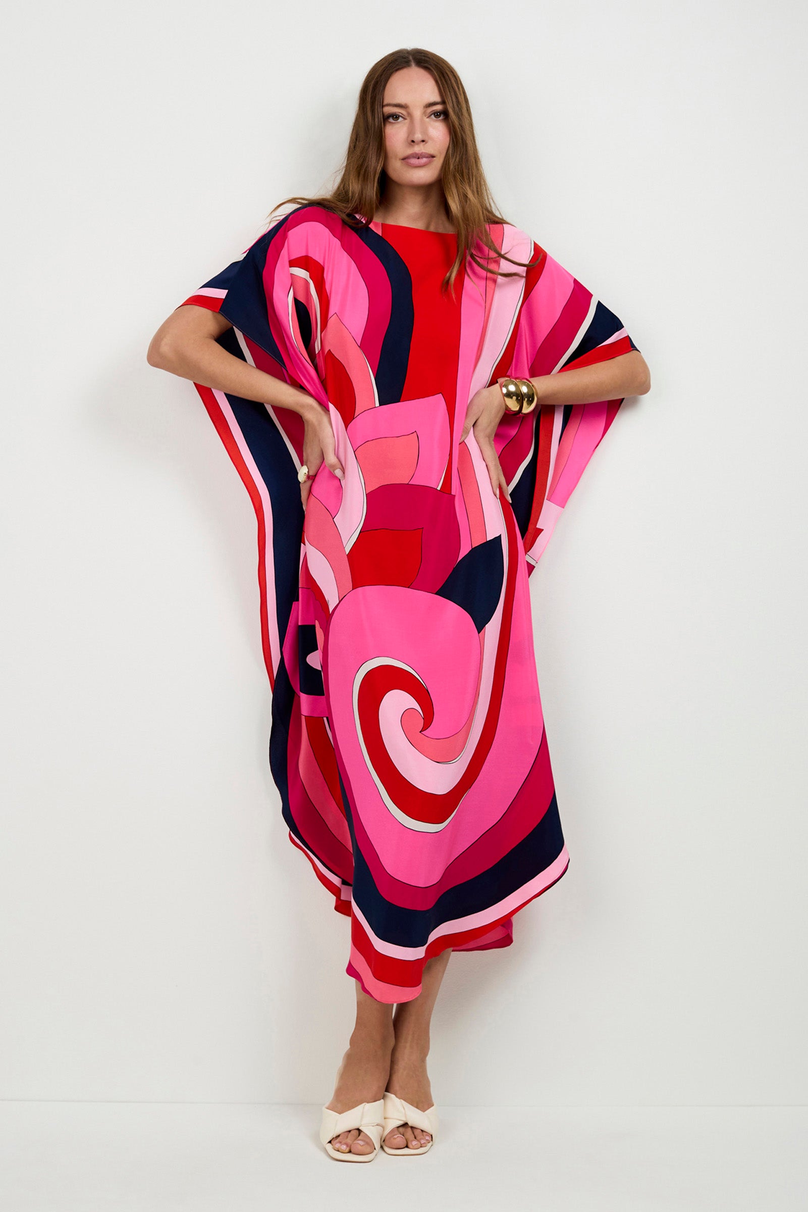 A woman stands against a white wall wearing the Jalani Maxi Caftan Dress, featuring bold pink, red, and navy swirling patterns, paired with white sandals and a gold bracelet.