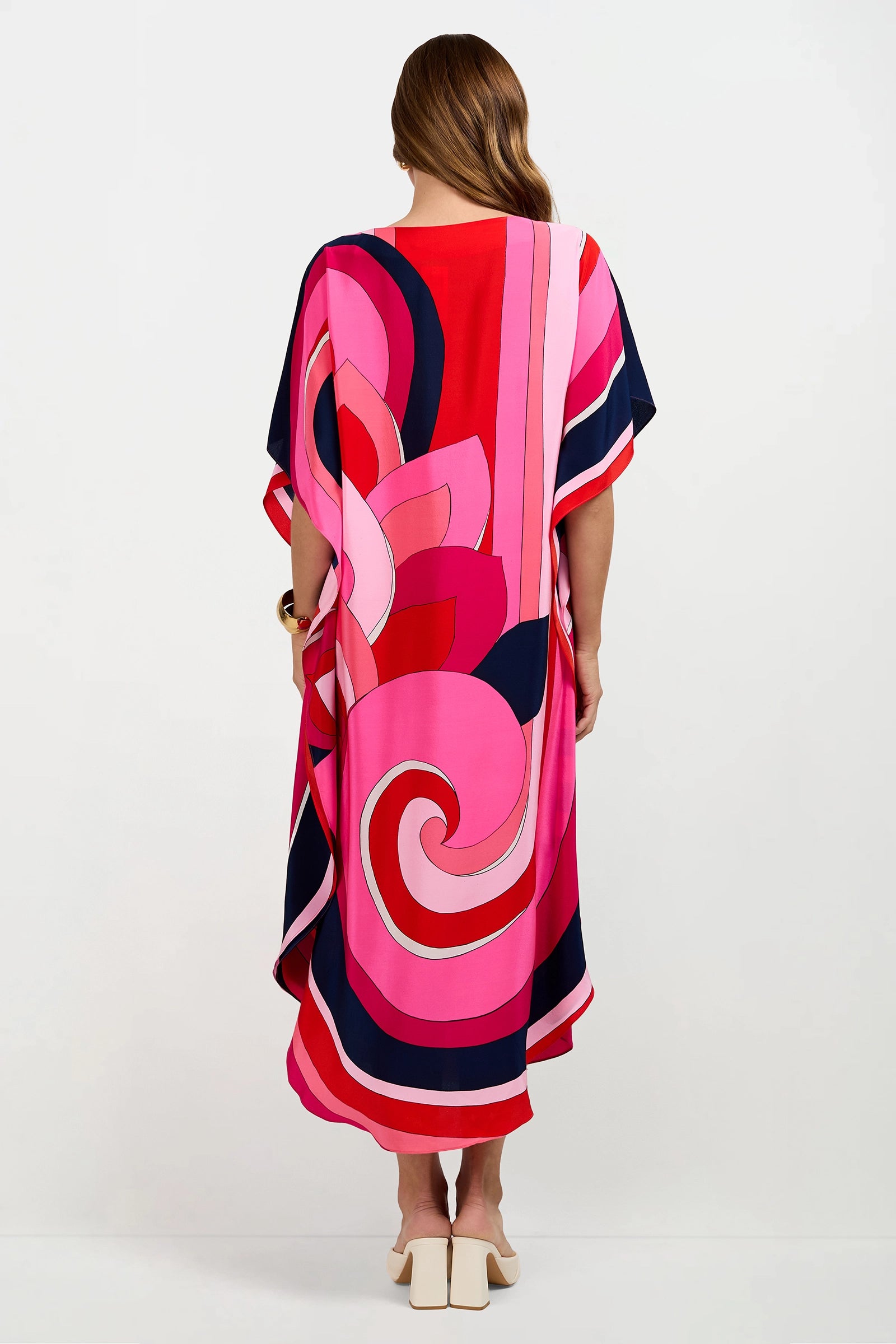 A woman with long brown hair faces away, wearing the Jalani Maxi Caftan Dress with bold pink, red, navy, and coral abstract patterns. She pairs it with white heeled sandals against a plain background.