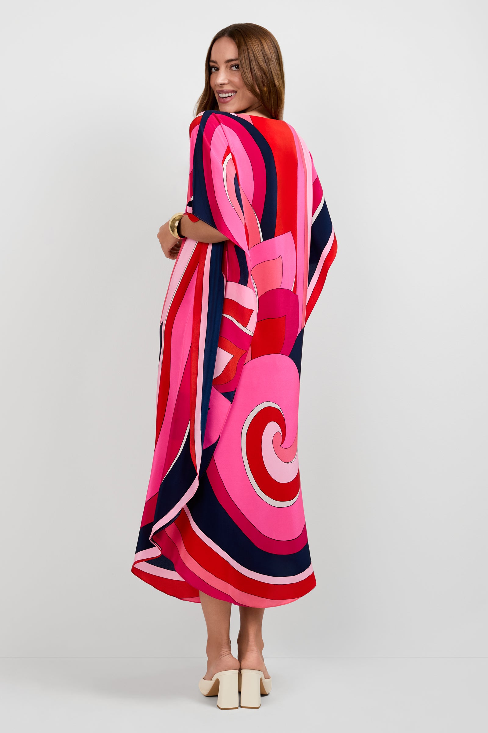 A woman with long brown hair smiles, turned to the side, wearing the Jalani Maxi Caftan Dress featuring bold pink, red, navy, and white swirl patterns. She completes the look with white heeled sandals.