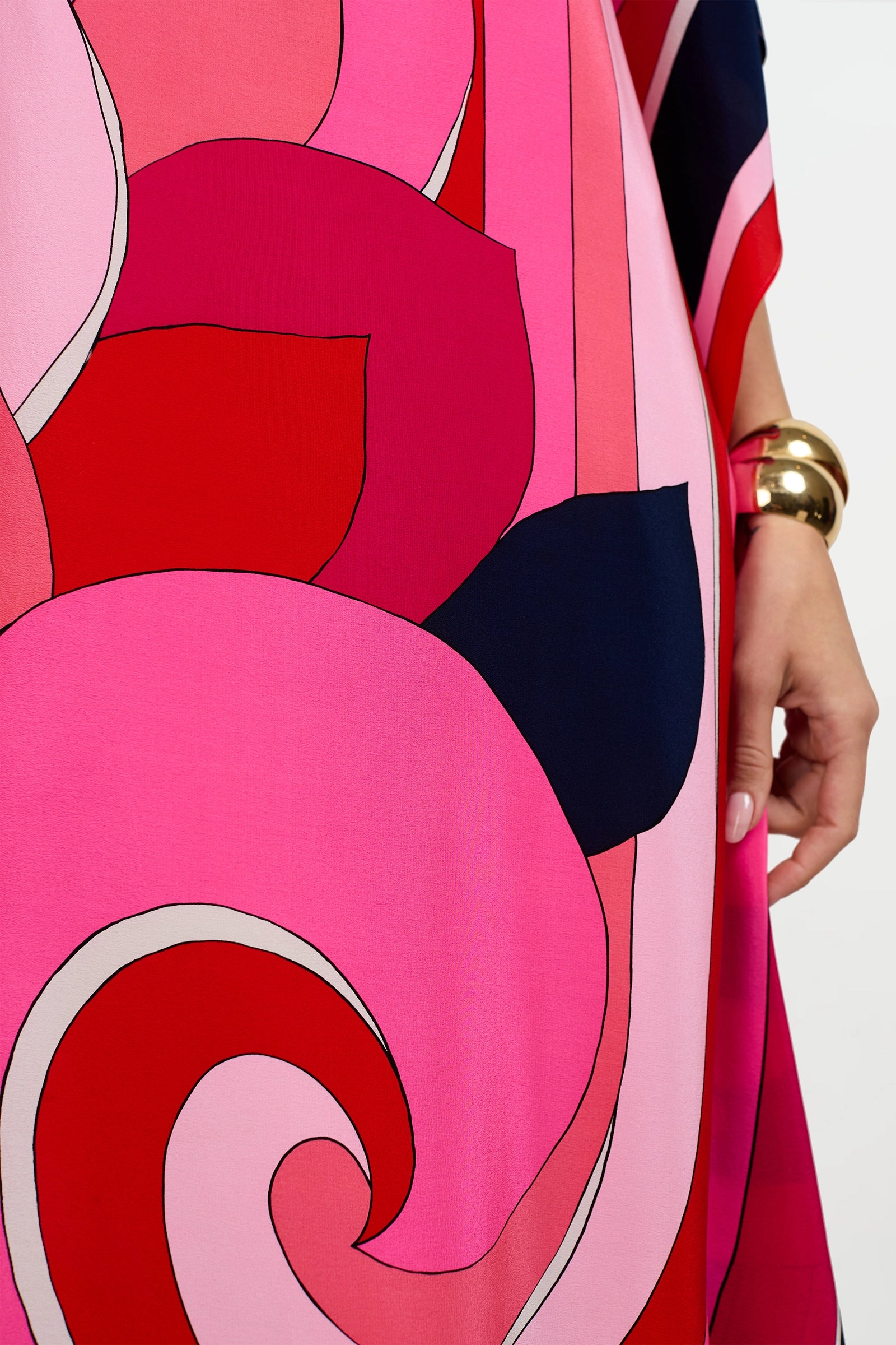 A lower arm wearing a gold bracelet is visible, along with part of the vibrant Jalani Maxi Caftan Dress featuring colorful abstract patterns in shades of pink, red, and navy.