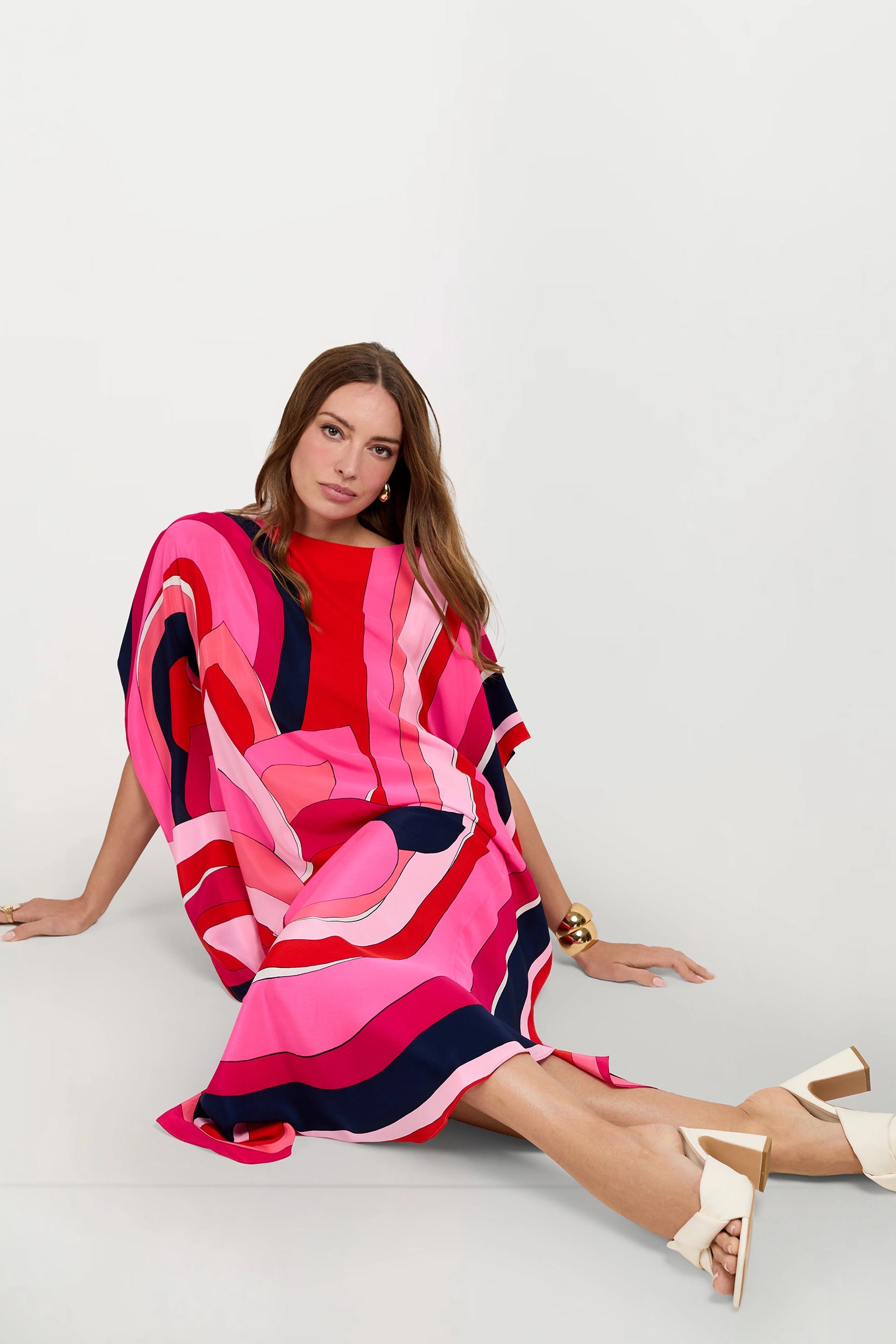 A woman with long brown hair sits on the floor against a white background, wearing the Jalani Maxi Caftan Dress featuring bold pink, red, and navy abstract patterns, paired with white sandal heels.