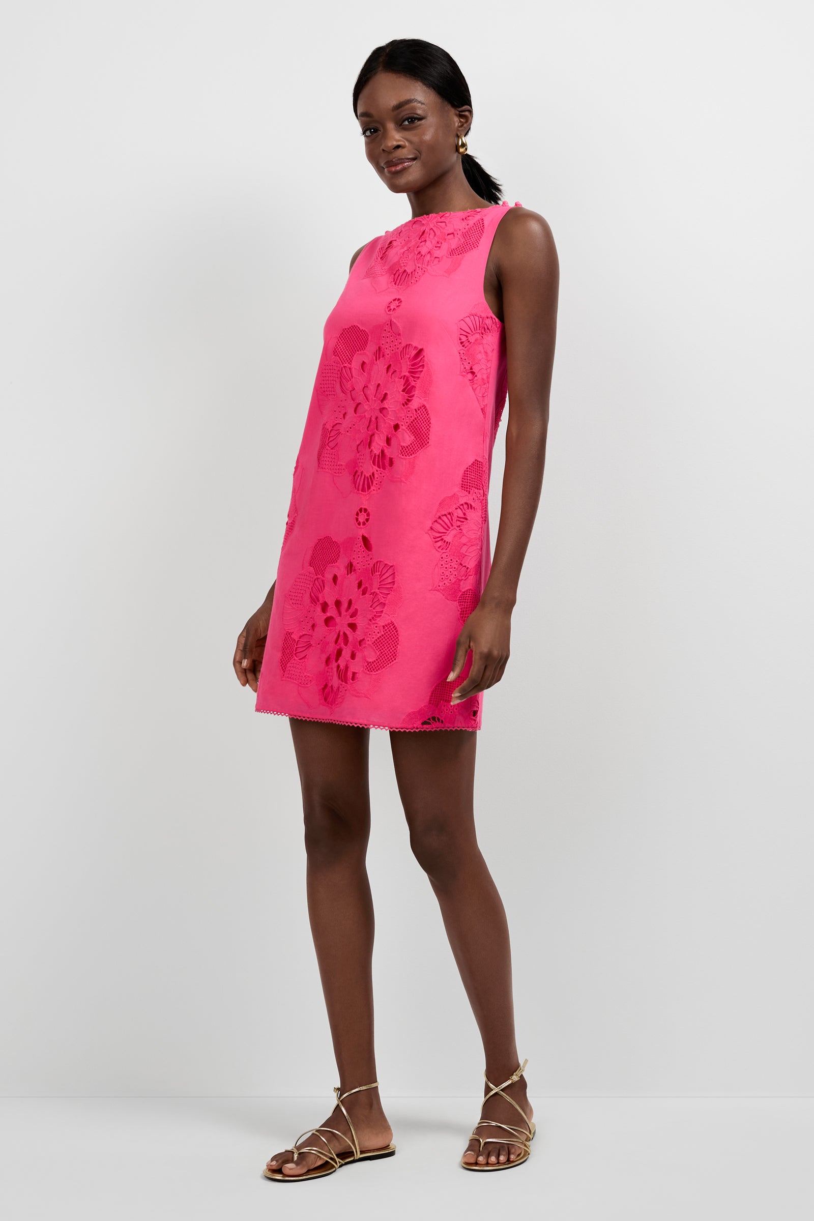 A woman stands against a plain background wearing the Francesca Shift Dress, a sleeveless bright pink mini dress with large floral patterns, paired with gold strappy flat sandals. Her hair is tied back and she smiles slightly.