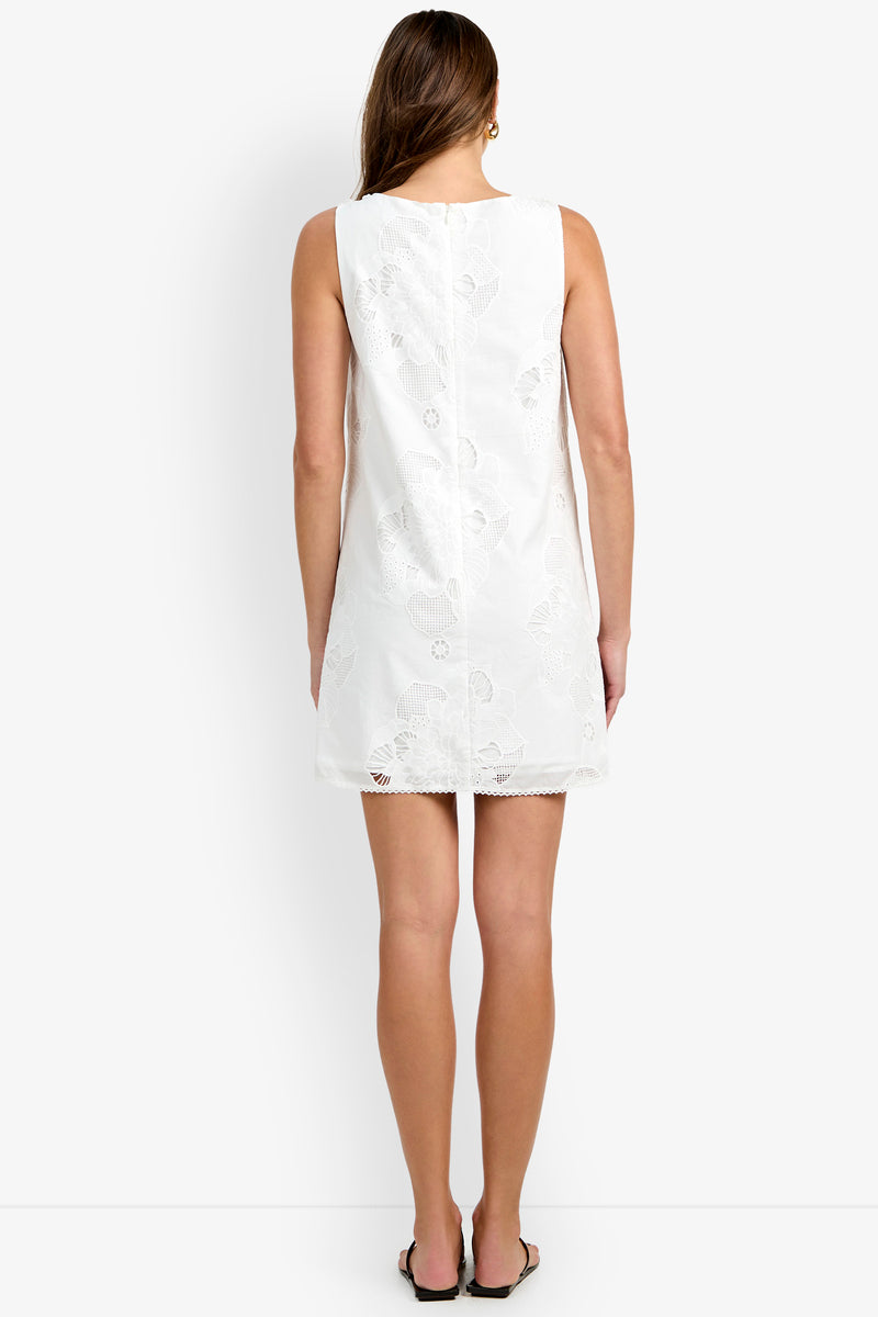 A woman stands facing away, wearing the sleeveless Francesca Shift Dress—a short white dress with a subtle floral pattern—paired with flat sandals, against a plain white background.