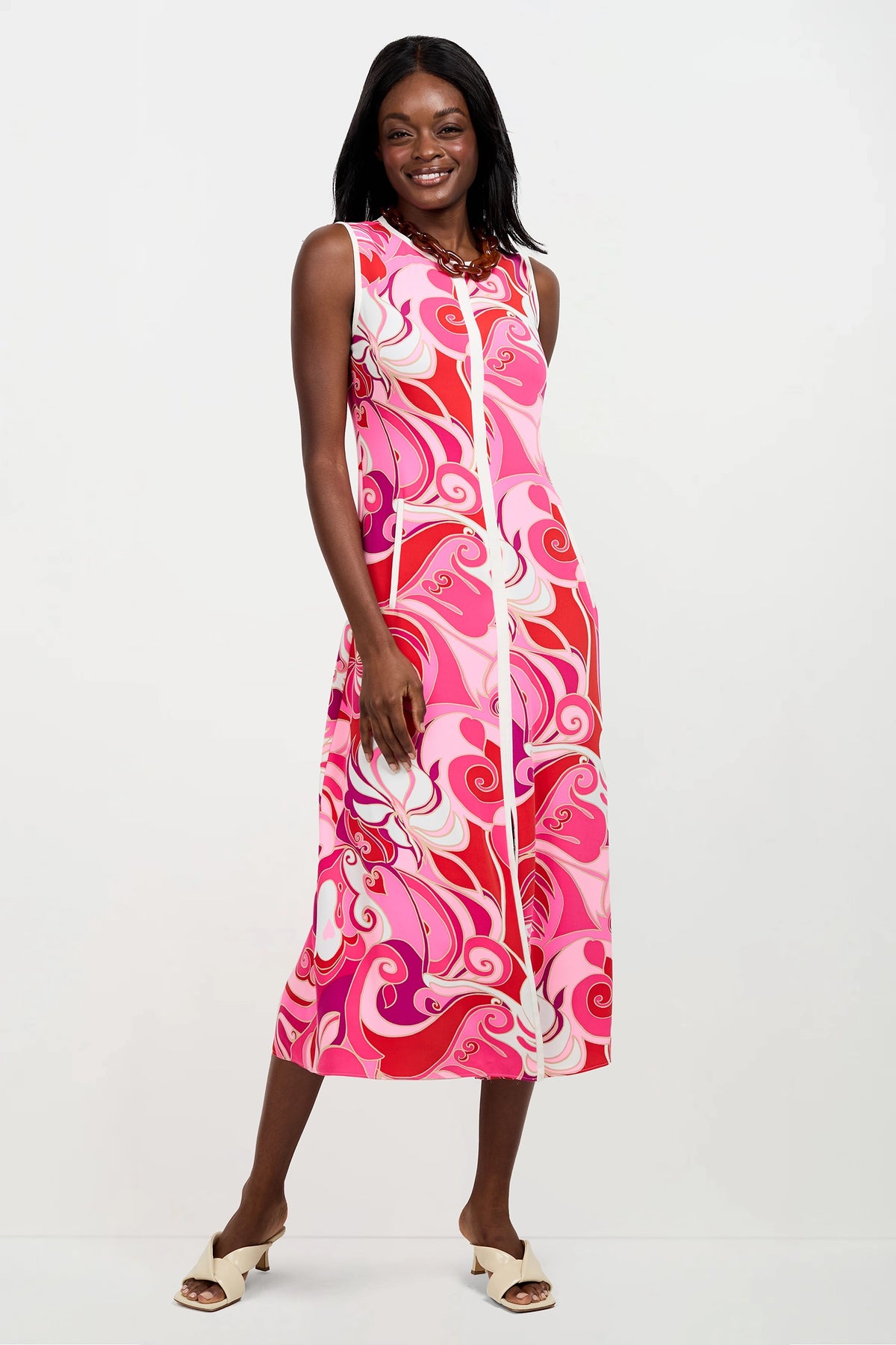 A woman smiles wearing the Magnifique Sleeveless Dress in Darsena, featuring a bold swirling pattern. She stands against a plain white background and completes her look with white open-toe heels.