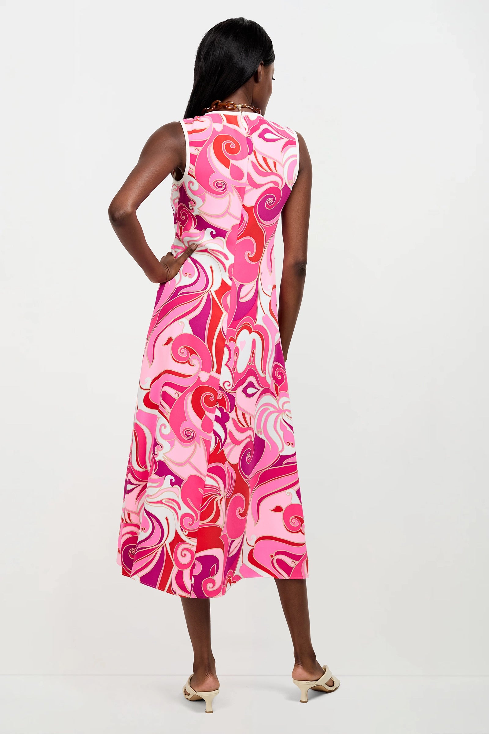 A woman with dark hair stands with her back to the camera, wearing the Magnifique Sleeveless Dress—a calf-length piece featuring a vibrant pink, red, and white swirling pattern—paired with heeled sandals against a plain white background.
