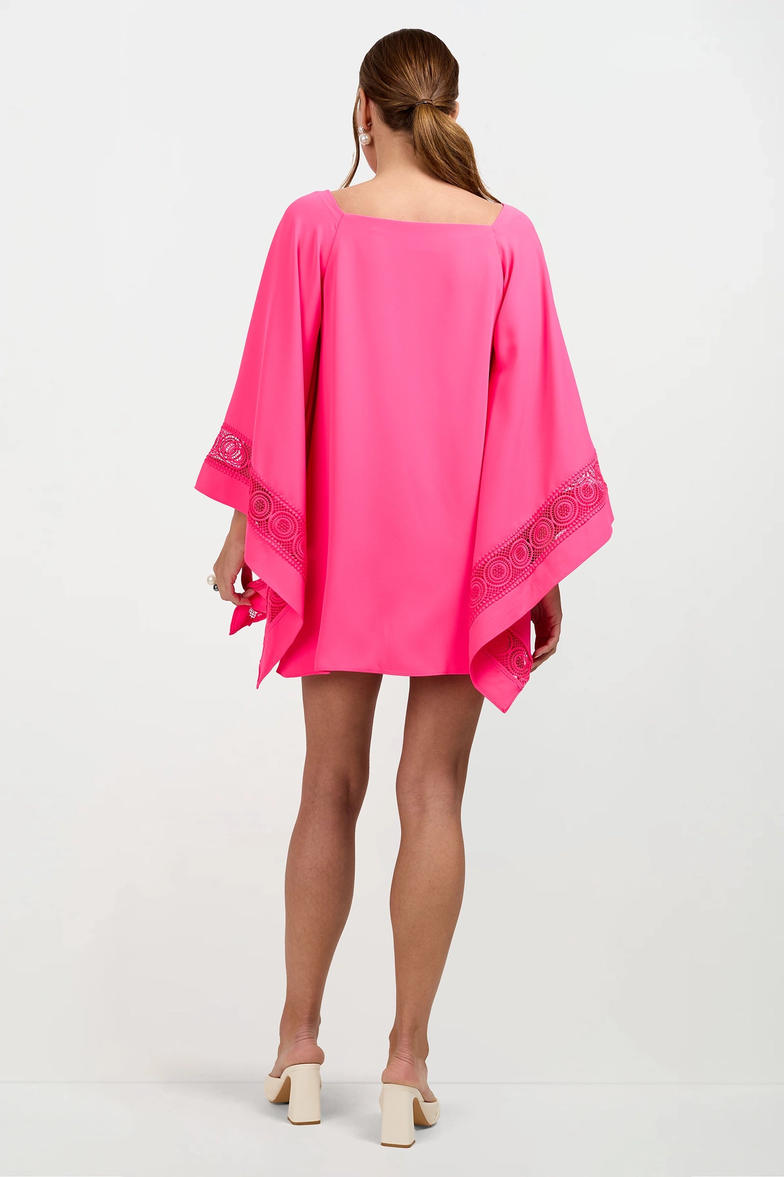 A woman with long brown hair tied back wears the Aqualane Side Flounce Dress, a loose, bright pink style with wide sleeves and decorative trim. She pairs it with light block-heeled shoes against a white background.