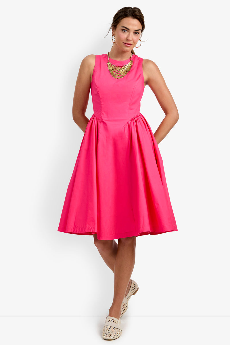 A woman stands against a plain white background wearing the Lombardy Midi Dress, a sleeveless, knee-length bright pink dress with pockets. She accessorizes with a chunky gold necklace and light-colored slip-on shoes.