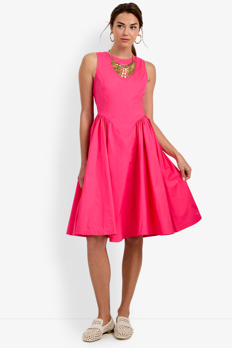 A woman wearing the Lombardy Midi Dress, which is sleeveless and bright pink with a knee-length hem, stands against a plain white background. She accessorizes with a chunky gold necklace and white woven slip-on shoes, holding part of her dress in one hand.