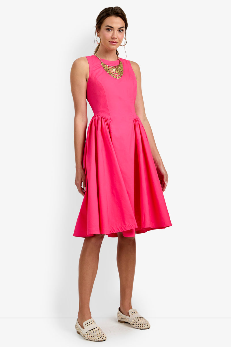 Woman wearing the Lombardy Midi Dress—a sleeveless, knee-length, bright pink dress with a flared skirt—paired with beige woven shoes and a chunky necklace, standing against a plain white background.