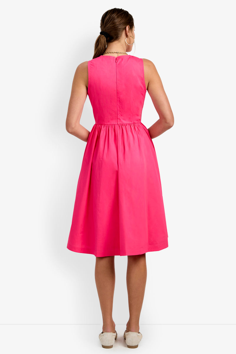A woman stands facing away, wearing the Lombardy Midi Dress—a sleeveless, knee-length bright pink dress with a gathered waist. She has light brown hair styled in a low ponytail and wears white shoes, with her hands behind her back.