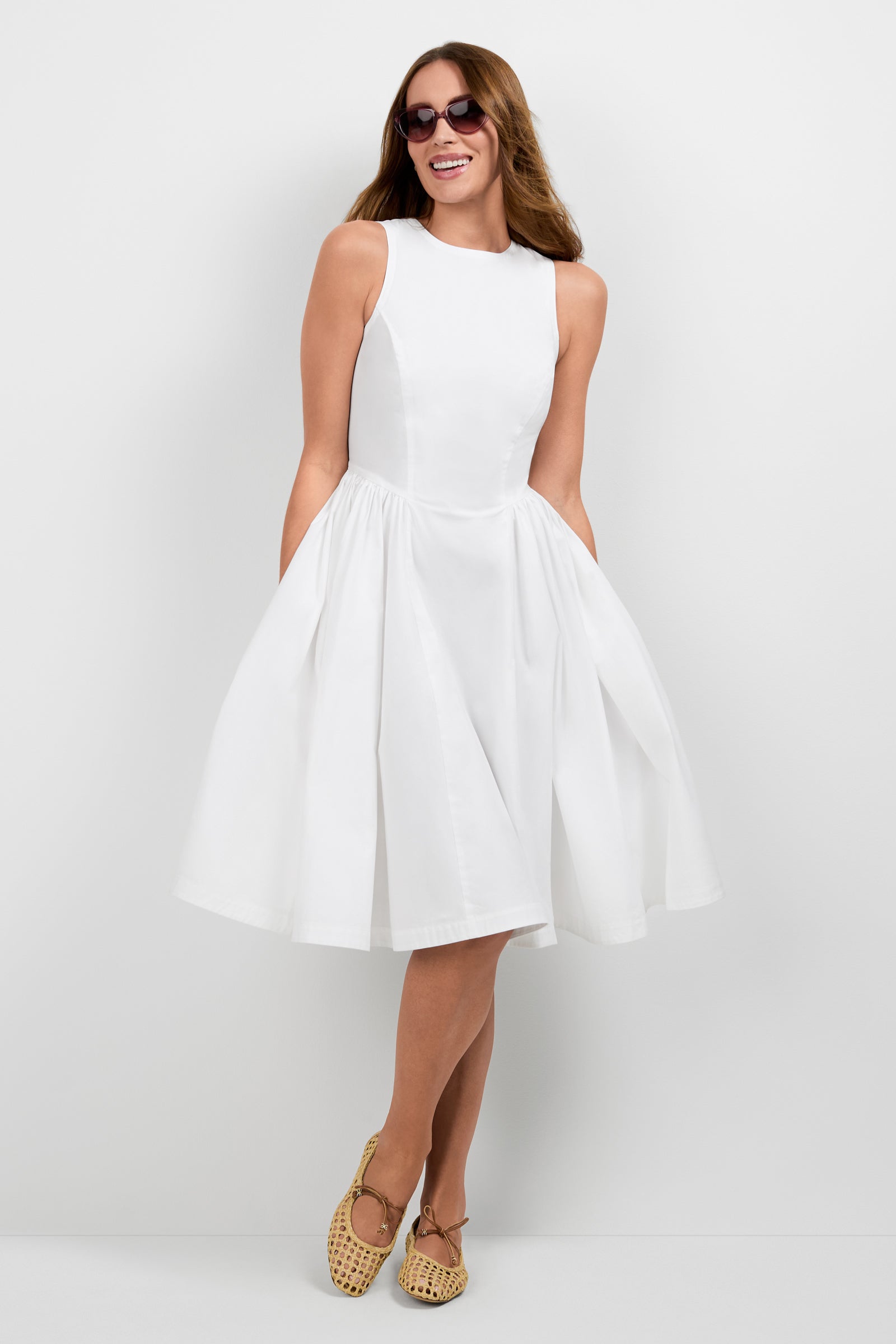 A smiling woman stands against a plain white background wearing sunglasses, beige woven shoes, and the Lombardy Midi Dress—sleeveless, white, knee-length, and with a flared skirt.