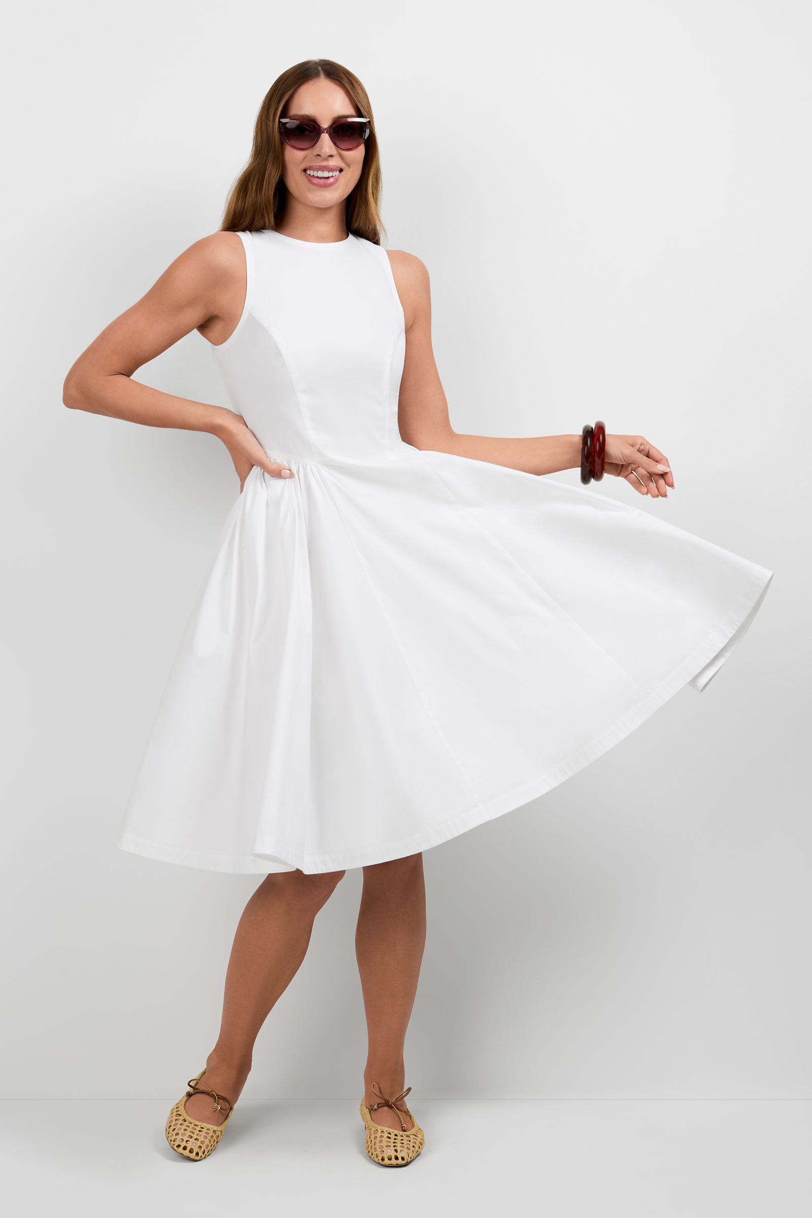 A smiling woman with long hair and sunglasses poses playfully in the sleeveless, knee-length Lombardy Midi Dress, holding the skirt to one side. She accessorizes with flat woven shoes and a red bangle against a plain white background.