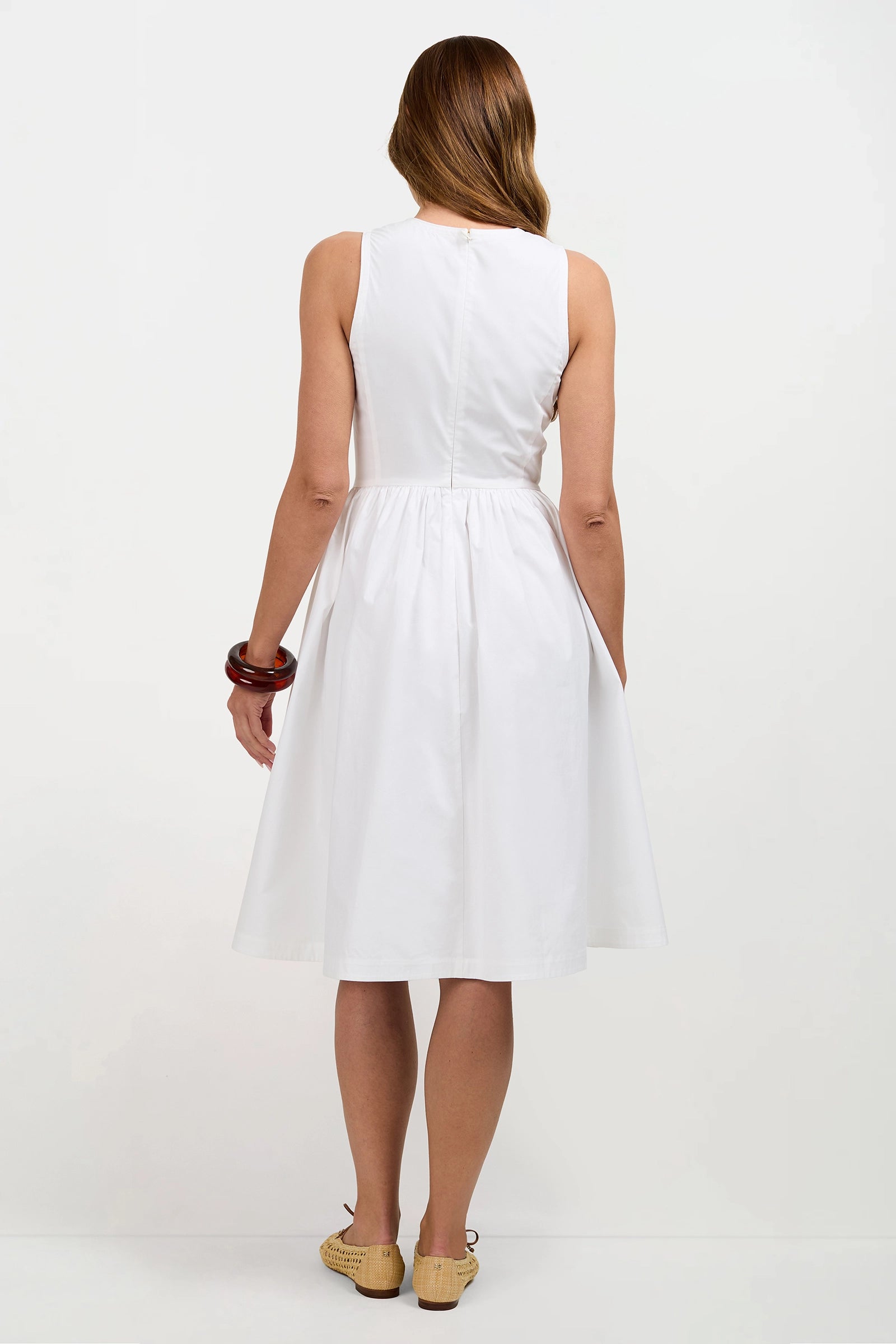 Woman in the Lombardy Midi Dress, a sleeveless white knee-length style, stands with her back to the camera against a white background. She pairs it with tan woven shoes, a brown bangle on her right wrist, and has long light brown hair.