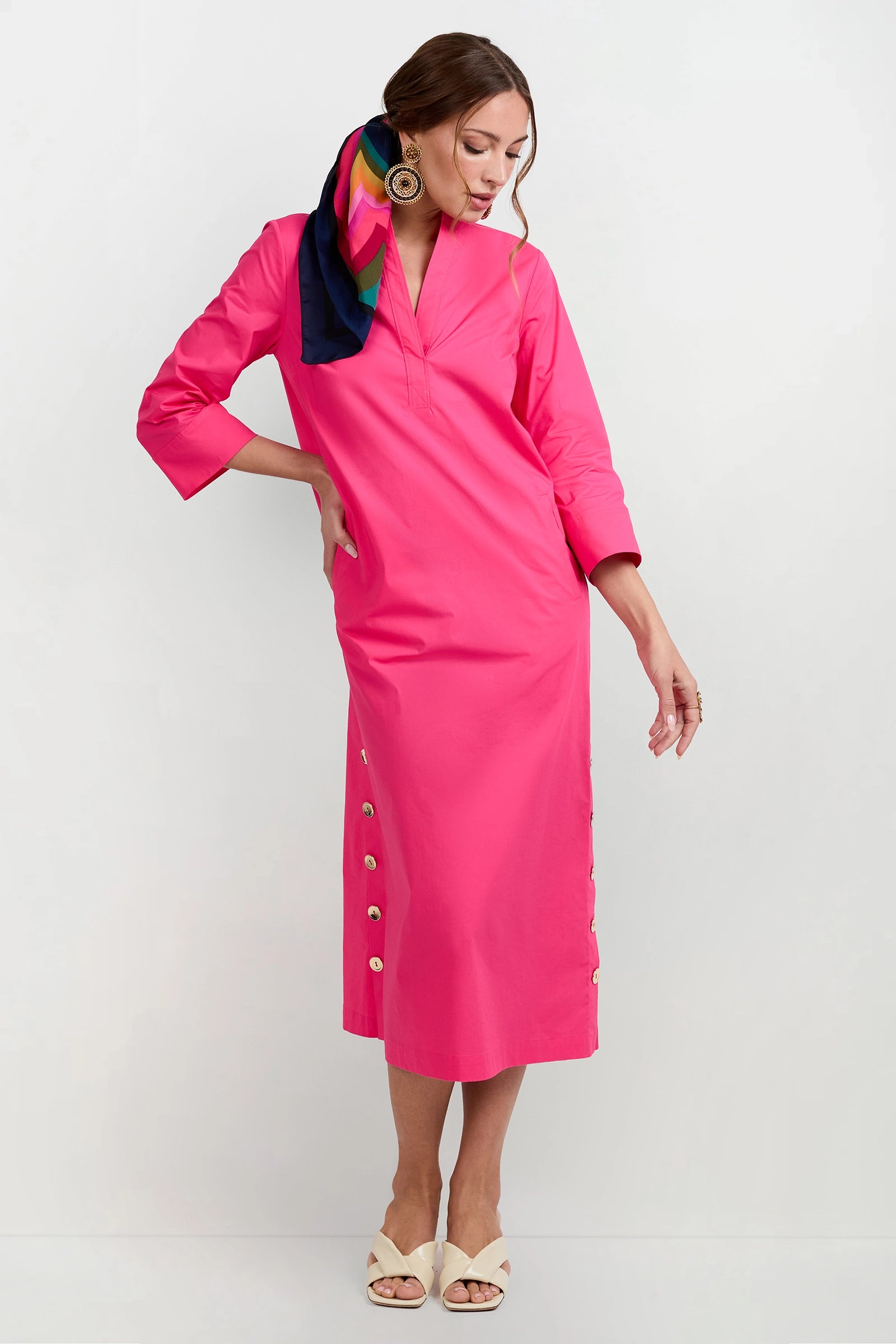A woman poses against a white background wearing the Capella Mandarin Collar Maxi Dress in bright pink, paired with cream sandals and a colorful scarf in her hair.