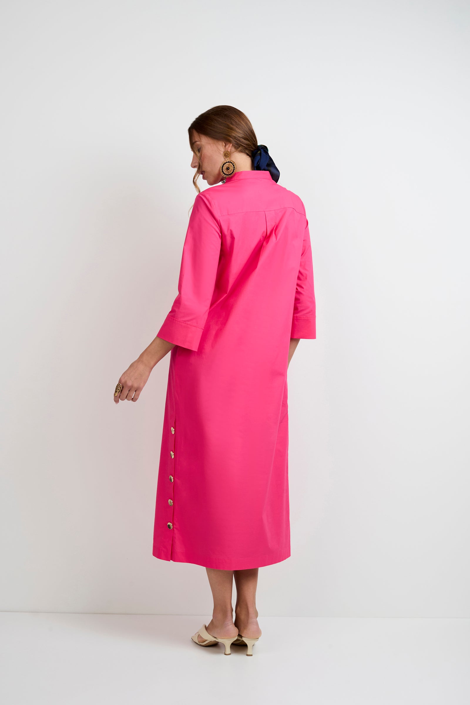 A woman with a navy scarf in her hair wears the Capella Mandarin Collar Maxi Dress in bright pink, featuring three-quarter sleeves and buttons, paired with beige open-toe heels, posing against a plain white background facing away.