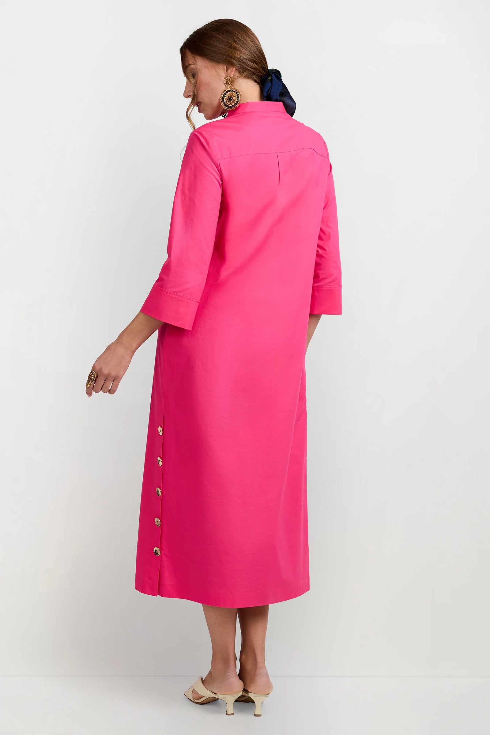 A woman models the Capella Mandarin Collar Maxi Dress in bright pink with side buttons, paired with beige heeled sandals and a dark hair ribbon, standing against a white background and looking over her shoulder.