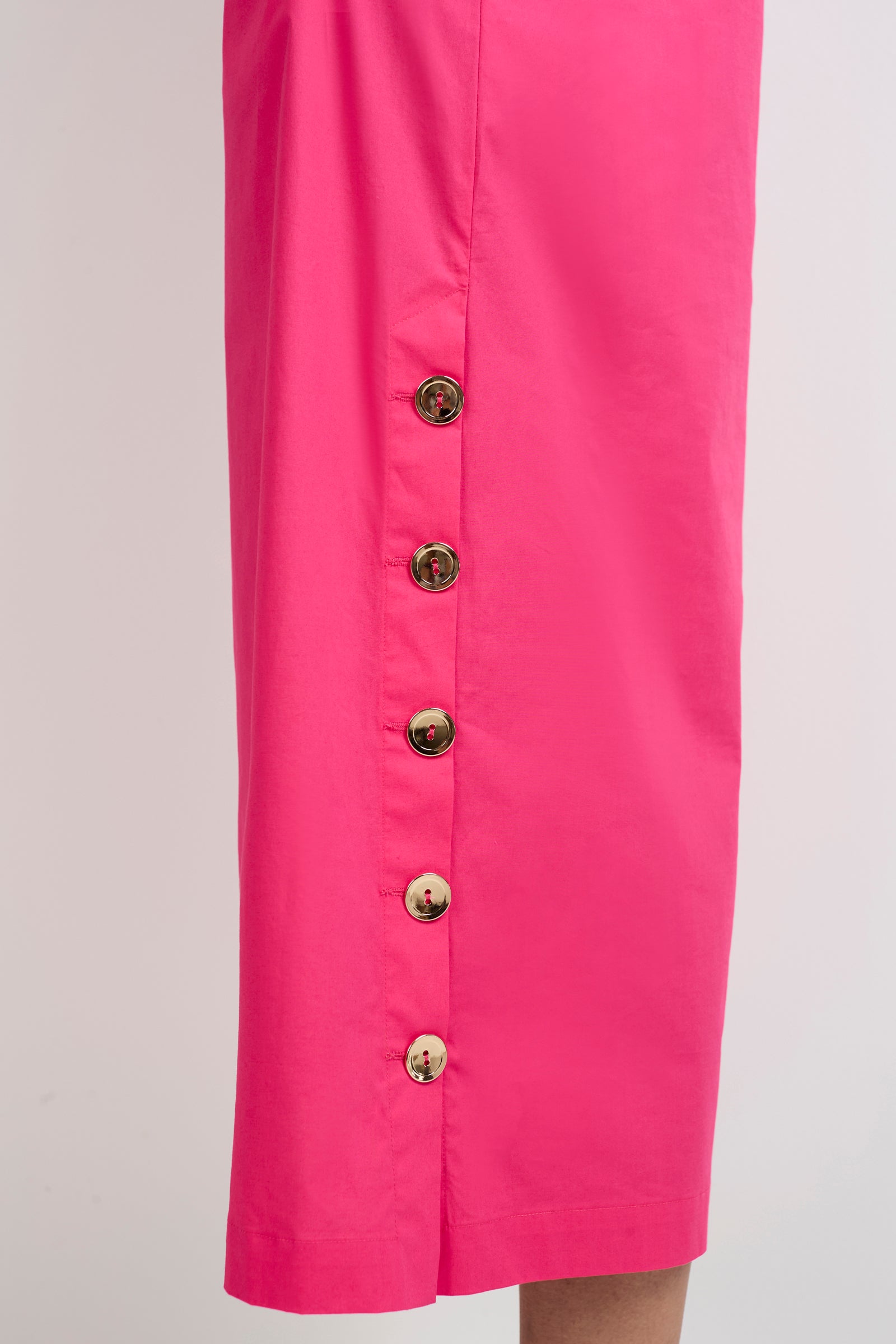A close-up of the Capella Mandarin Collar Maxi Dress in bright pink, showcasing six round gold buttons along the side hem. The dress fabric is smooth, set against a plain light-colored background.