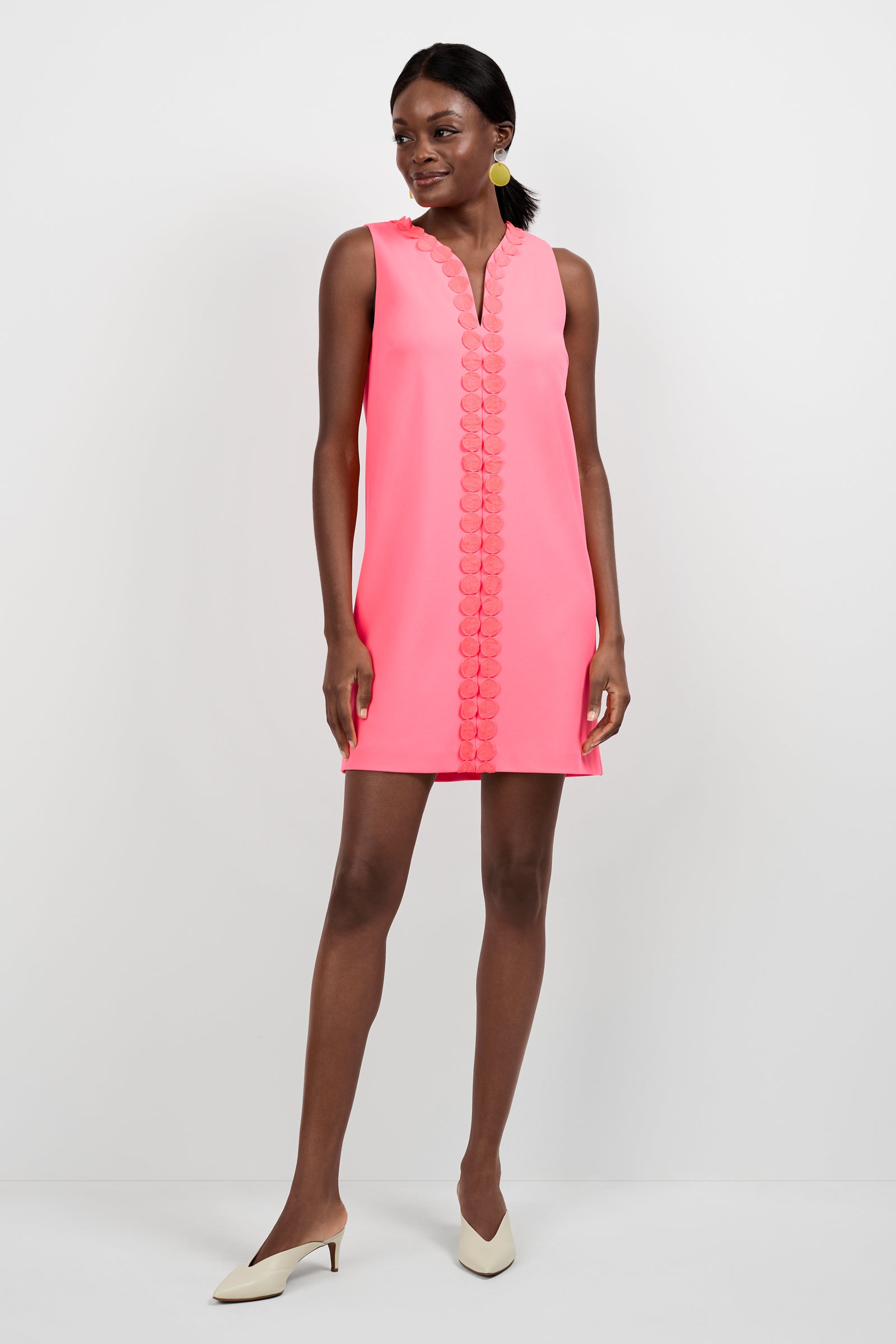 A woman smiles and looks away while wearing the Pavia Midi Dress—a sleeveless, bright pink shift dress with decorative front trim—paired with beige low-heeled shoes and yellow circular earrings, against a plain white background.