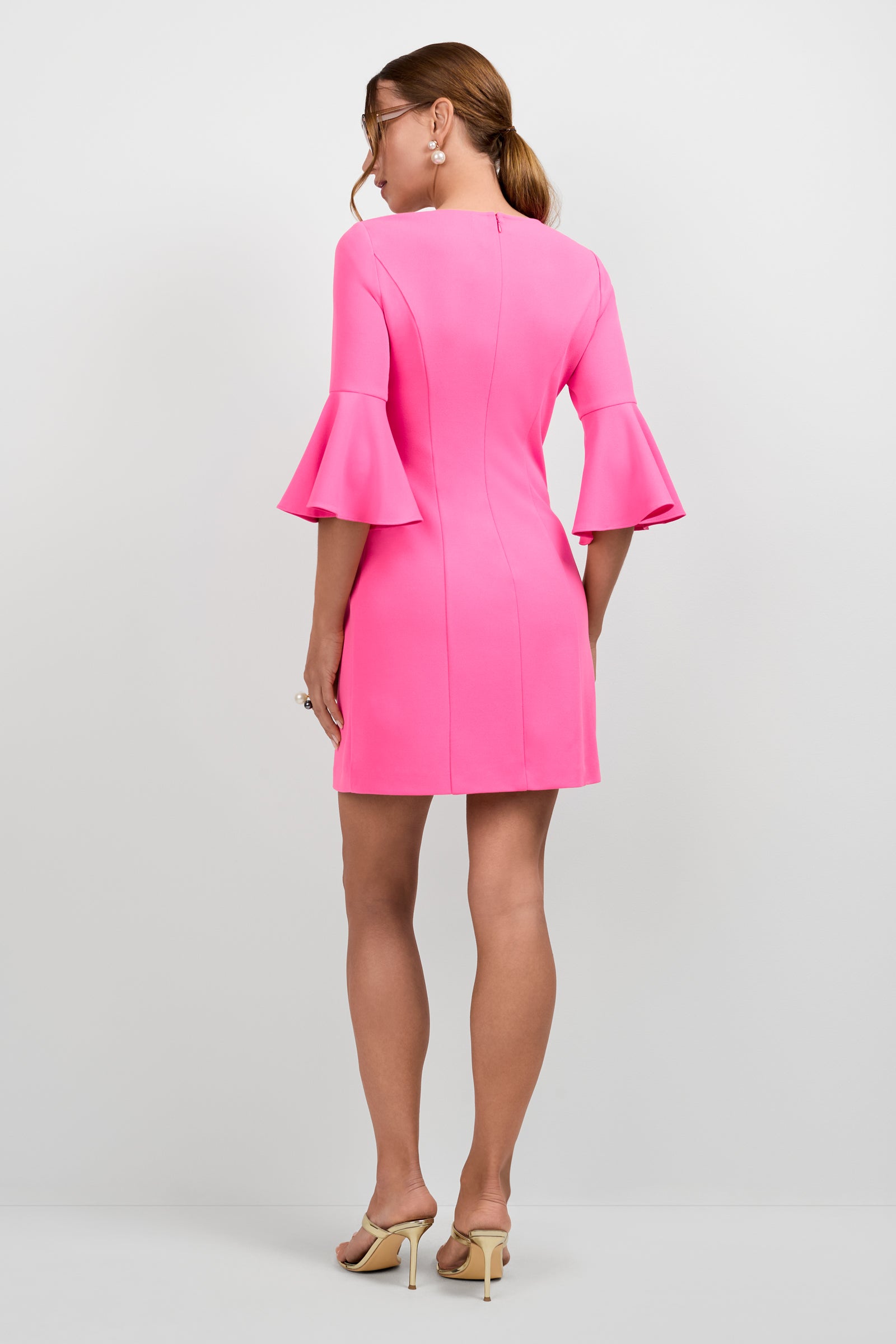A woman faces away in the Scala Flounce Sleeve Dress, a bright pink, knee-length style with ruffled three-quarter sleeves. She pairs it with nude high-heeled sandals, hoop earrings, and wears her light brown hair in a low ponytail.