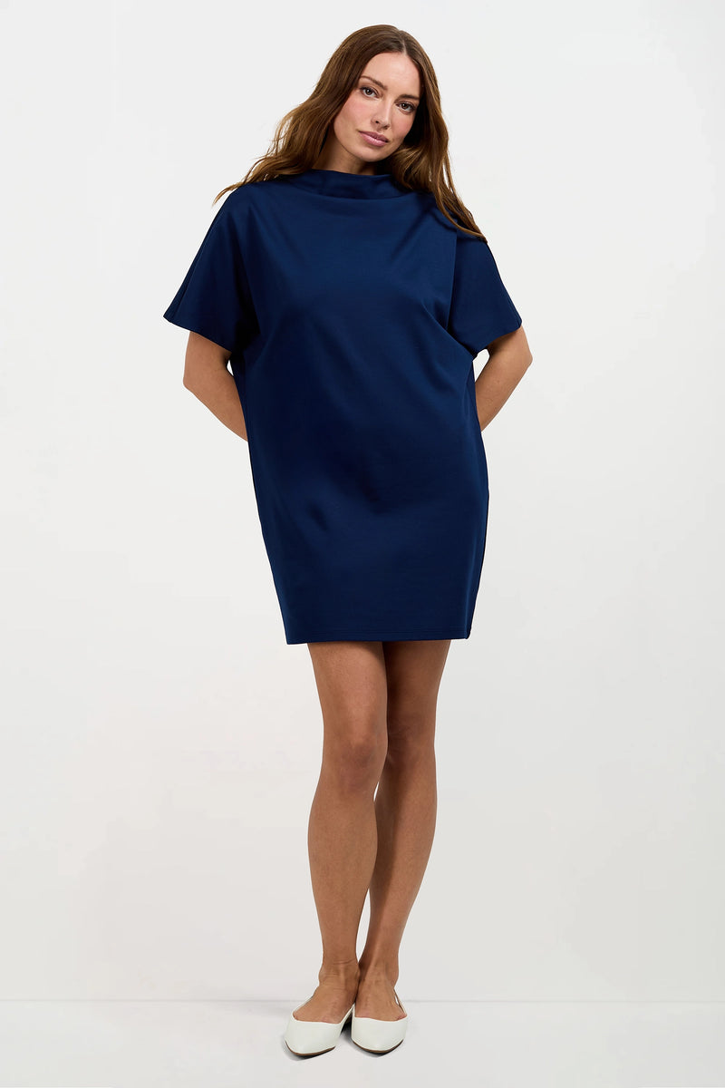 A woman with long brown hair and a relaxed expression stands against a white background, wearing the Storica Mini Dress—a short-sleeved, loose-fitting navy blue dress—with her hands behind her back and paired with white flats.