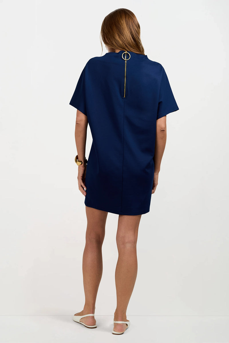 A woman with long brown hair stands with her back to the camera, wearing the Storica Mini Dress in dark blue featuring a gold zipper detail, paired with white flat shoes and a chunky bracelet.