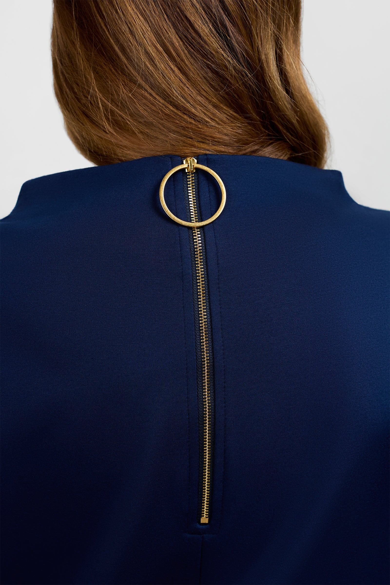 Close-up of the back of a person wearing the Storica Mini Dress in dark blue, featuring a visible gold zipper with a large circular gold pull. The person's brown hair is partially visible.