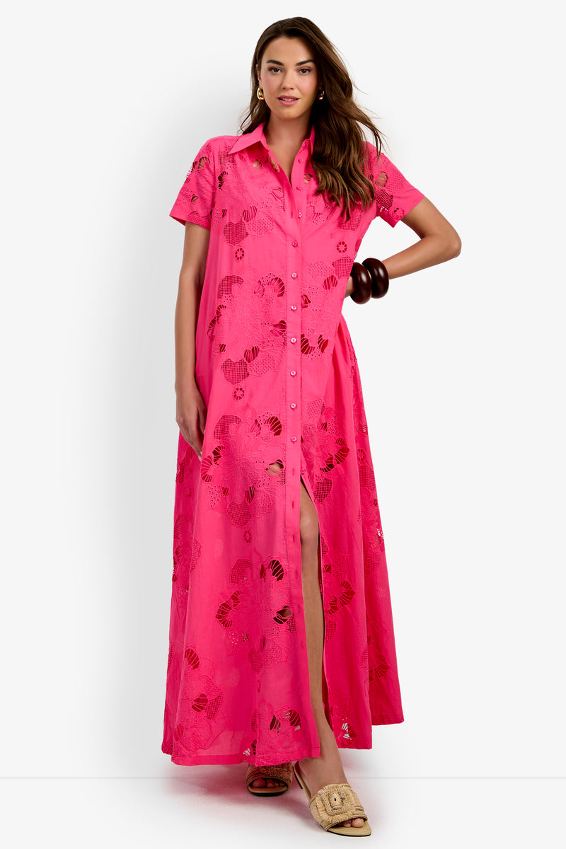 A woman stands against a white background wearing the Kon Tiki Maxi Dress—a bright pink button-up maxi dress featuring short sleeves, a collared neckline, and subtle floral patterns—accessorized with brown bangles and beige open-toe sandals.